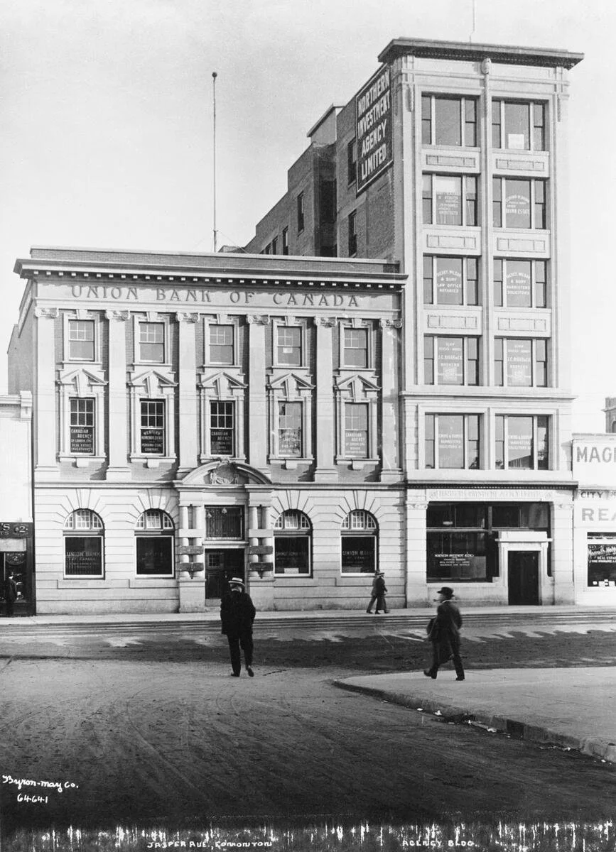 Historical black-and-white photo of a city street corner with buildings, including a bank named 'Union Bank of Canada' and an insurance agency. Pedestrians are walking on the sidewalk.