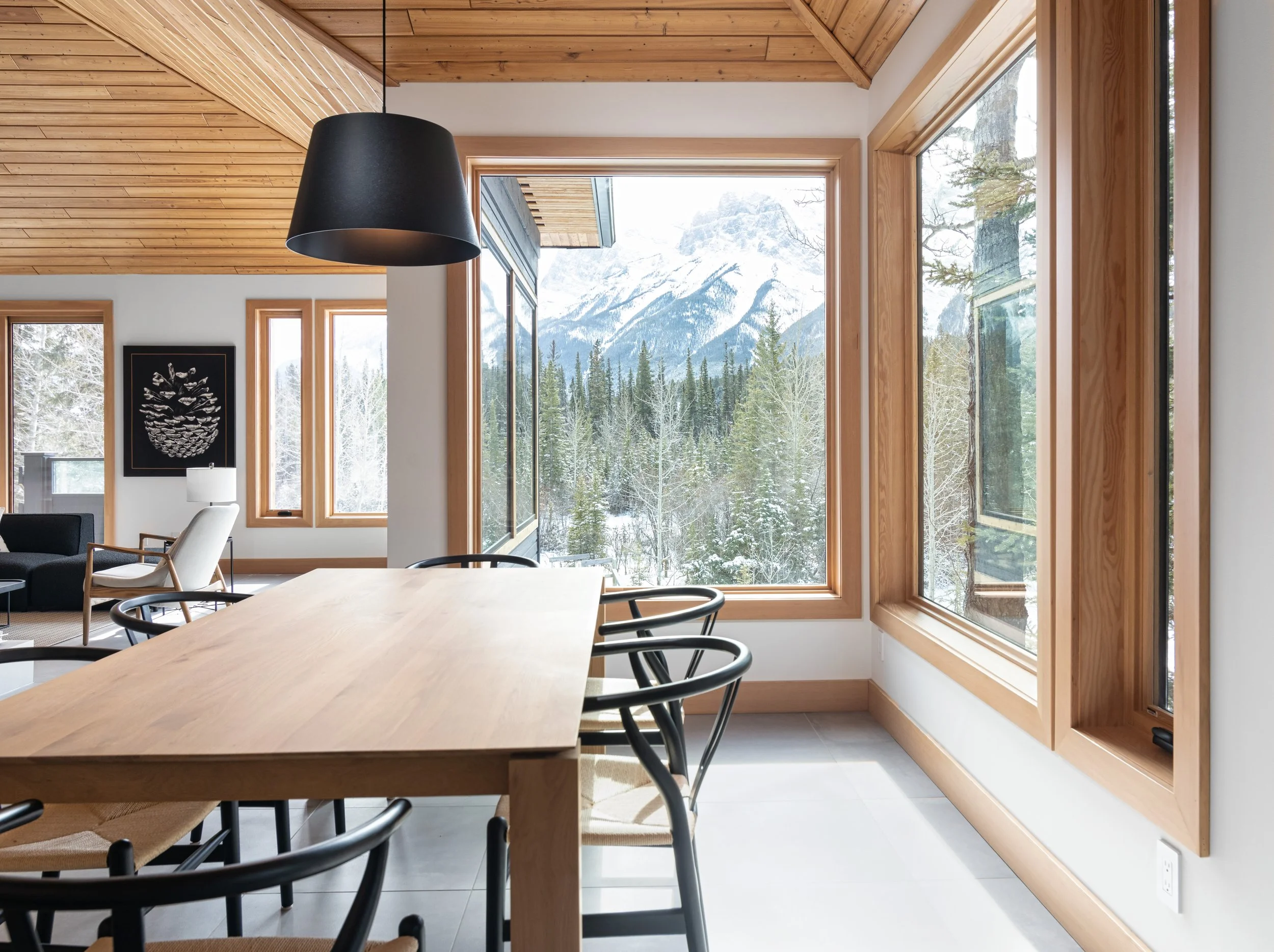 Interior view of a modern dining area with large windows showing a snowy mountain landscape, wooden ceiling, and table with black chairs.