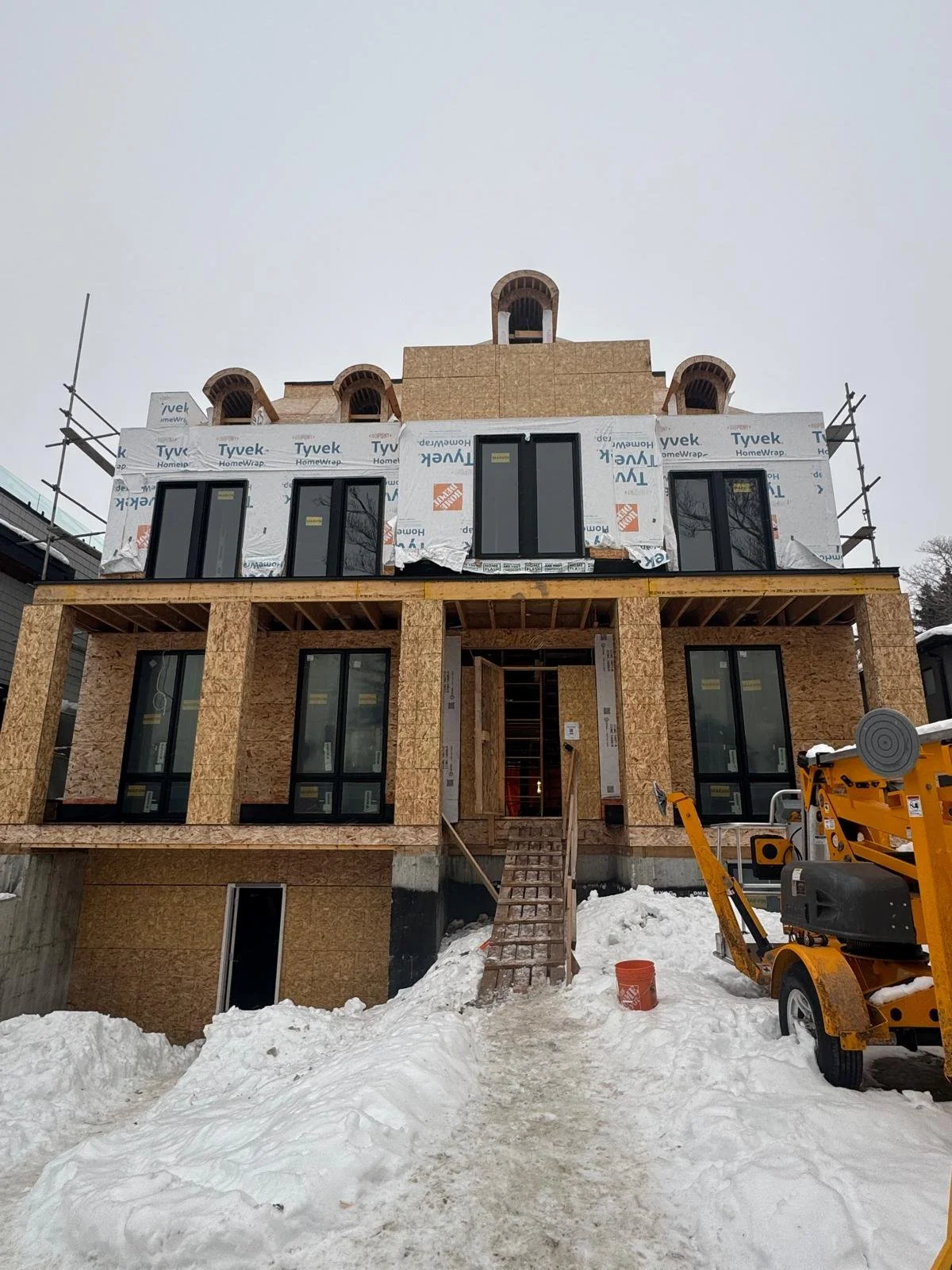 Under construction multi-story house with exposed wooden framing, black window frames, and snow on the ground, with construction equipment nearby.