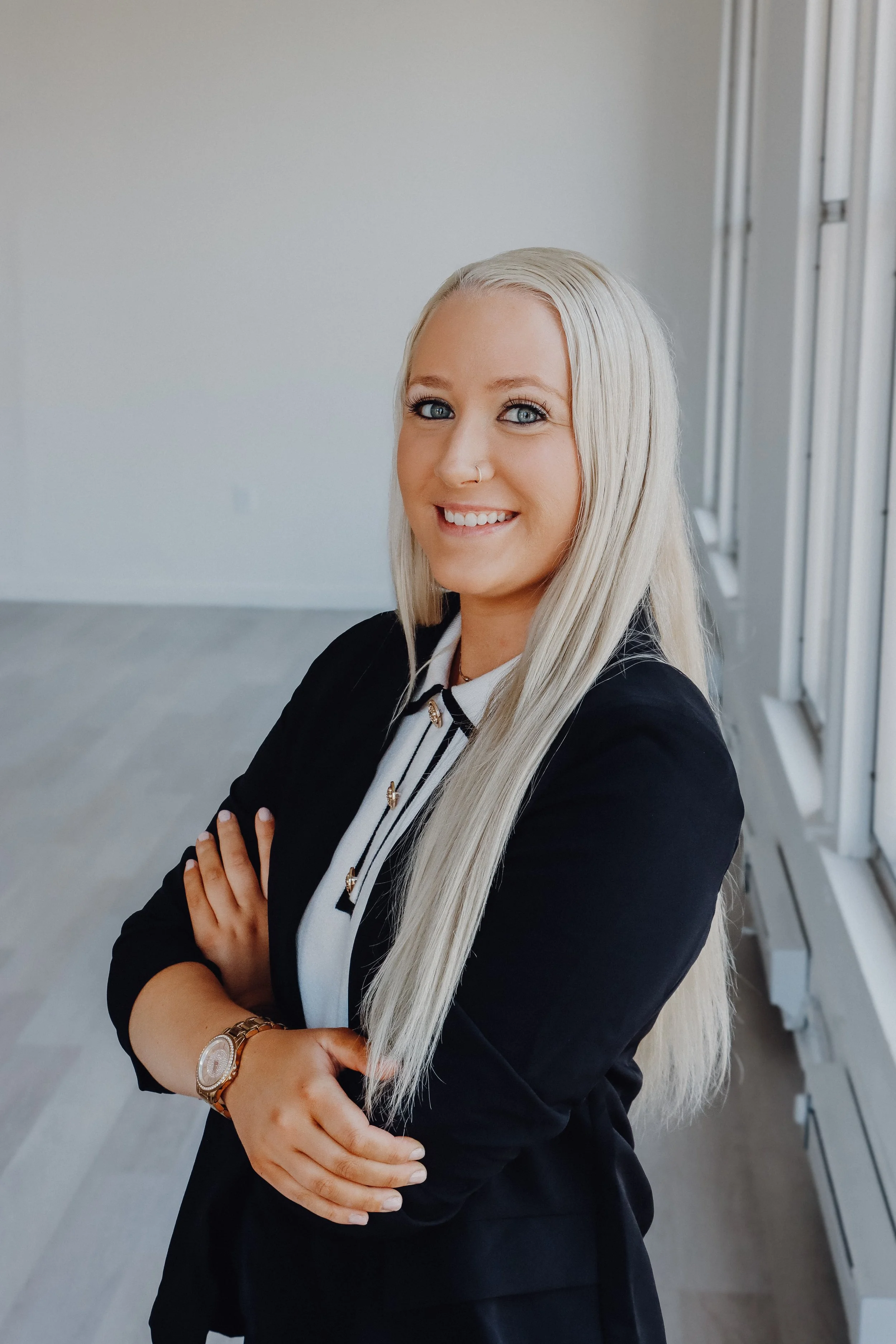 A woman with long blonde hair standing indoors, wearing a black blazer and white blouse, smiling with arms crossed. Modern office or conference room in the background.