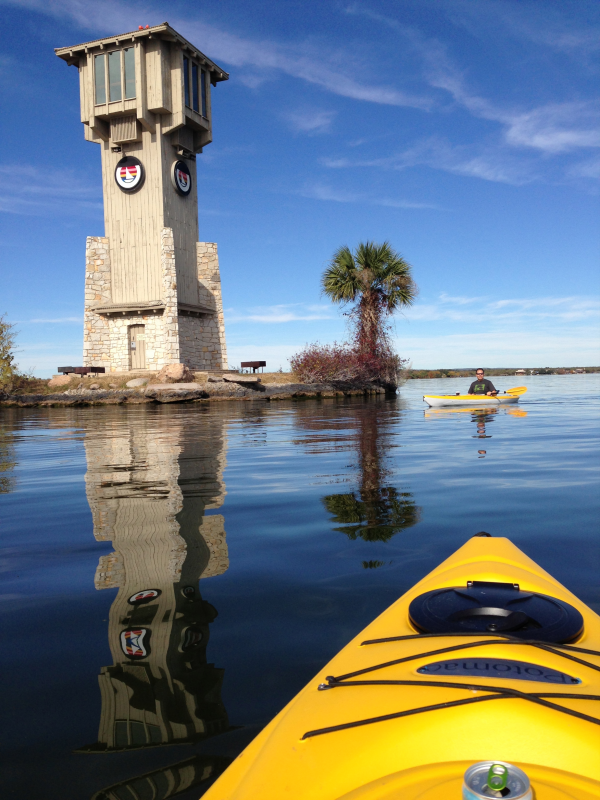 A kayaker on a calm body of water near a small island with a tall wooden observation tower, a palm tree, and two other kayakers in the distance.