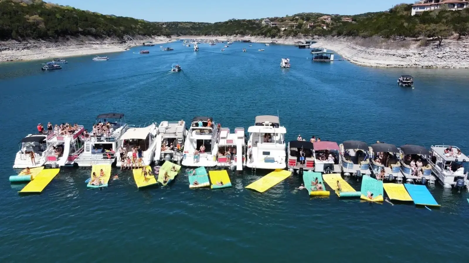 A marina with several boats docked and floating on the water, some people swimming and lounging on colorful float mats, with a background of hilly terrain and houses.