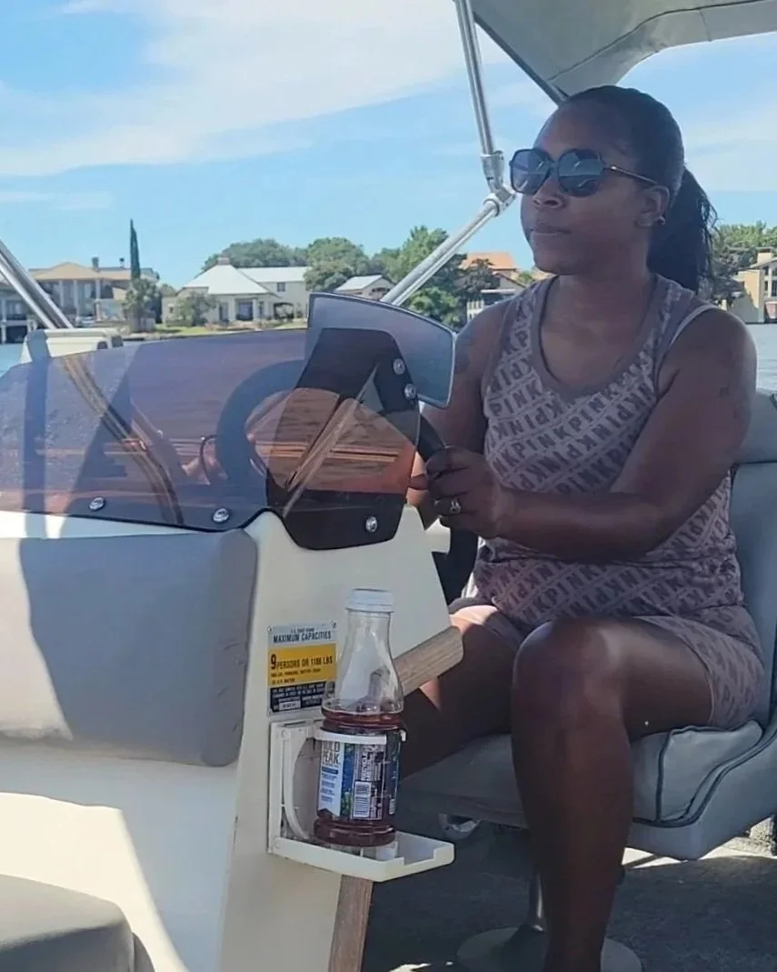 A woman wearing sunglasses and a tank top sits on a boat, holding a smartphone. There is a bottle of tea with a jar lid in a cup holder attached to the boat's console. The background shows houses and trees near a body of water under a partly cloudy sky.