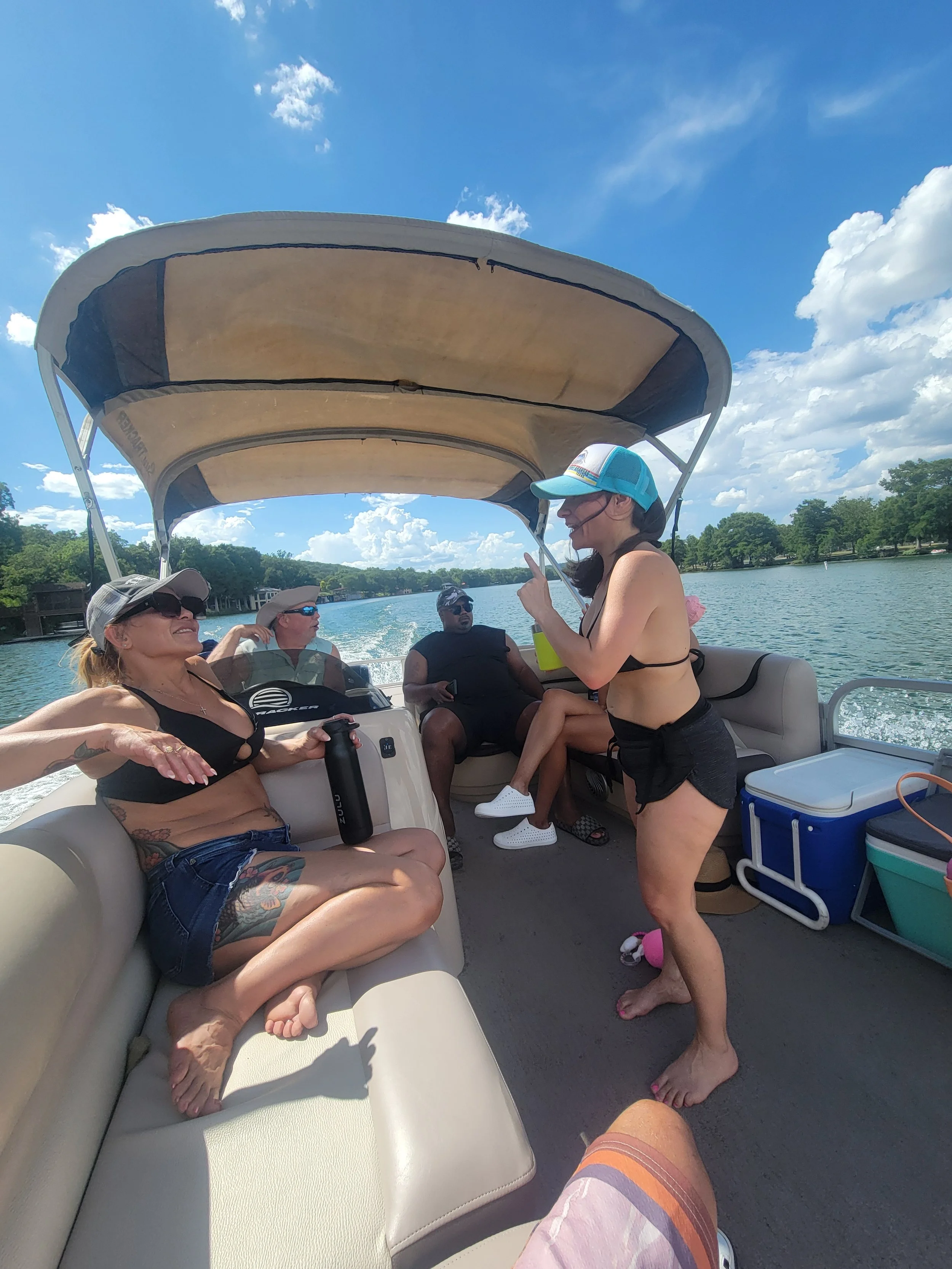 People relaxing and socializing on a boat on a lake under a sunny sky with clouds, some wearing hats and sunglasses.