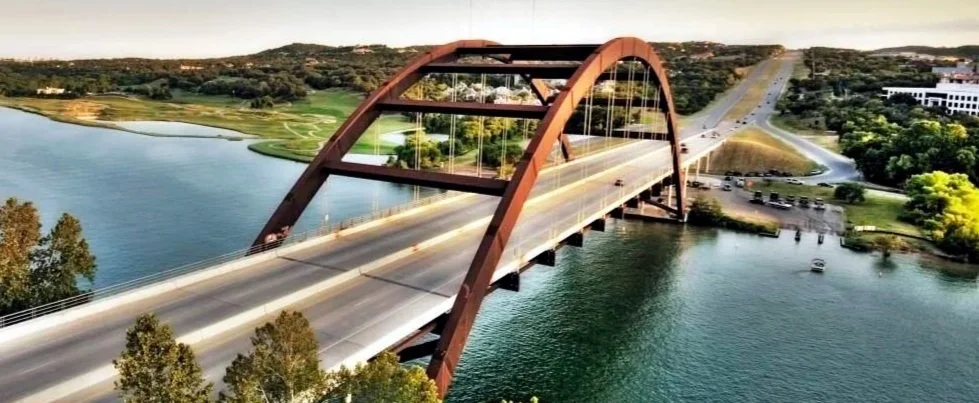 A beautiful overhead view of 360 Bridge in Austin, Texas spanning over a body of water, with trees and a parking lot nearby, under a clear sky.