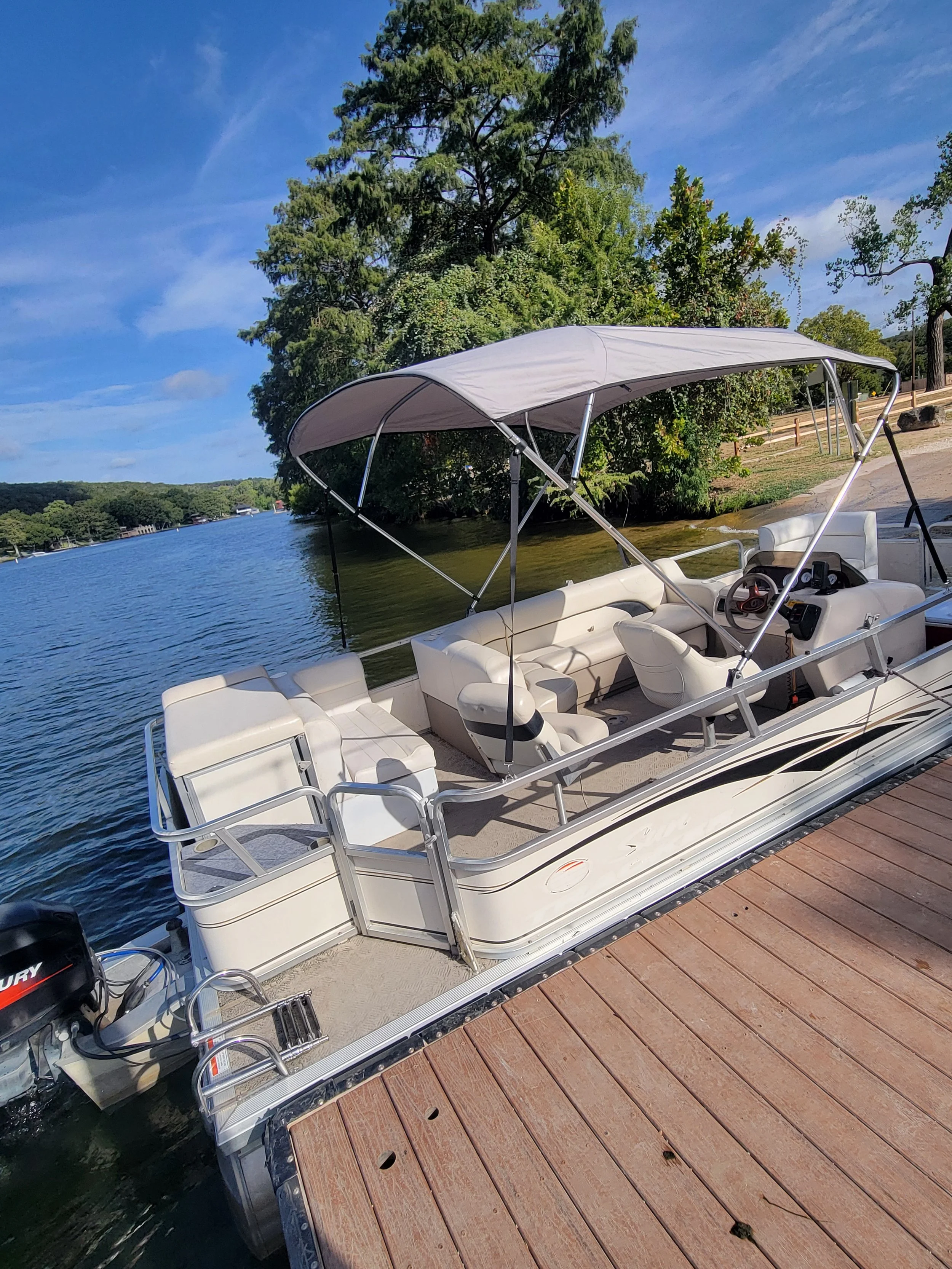 A white pontoon boat docked at a wooden pier on a lake, with trees and blue sky in the background.