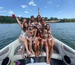 Group of nine people on a boat, wearing swimsuits, enjoying a sunny day on the water with trees and a clear sky in the background.