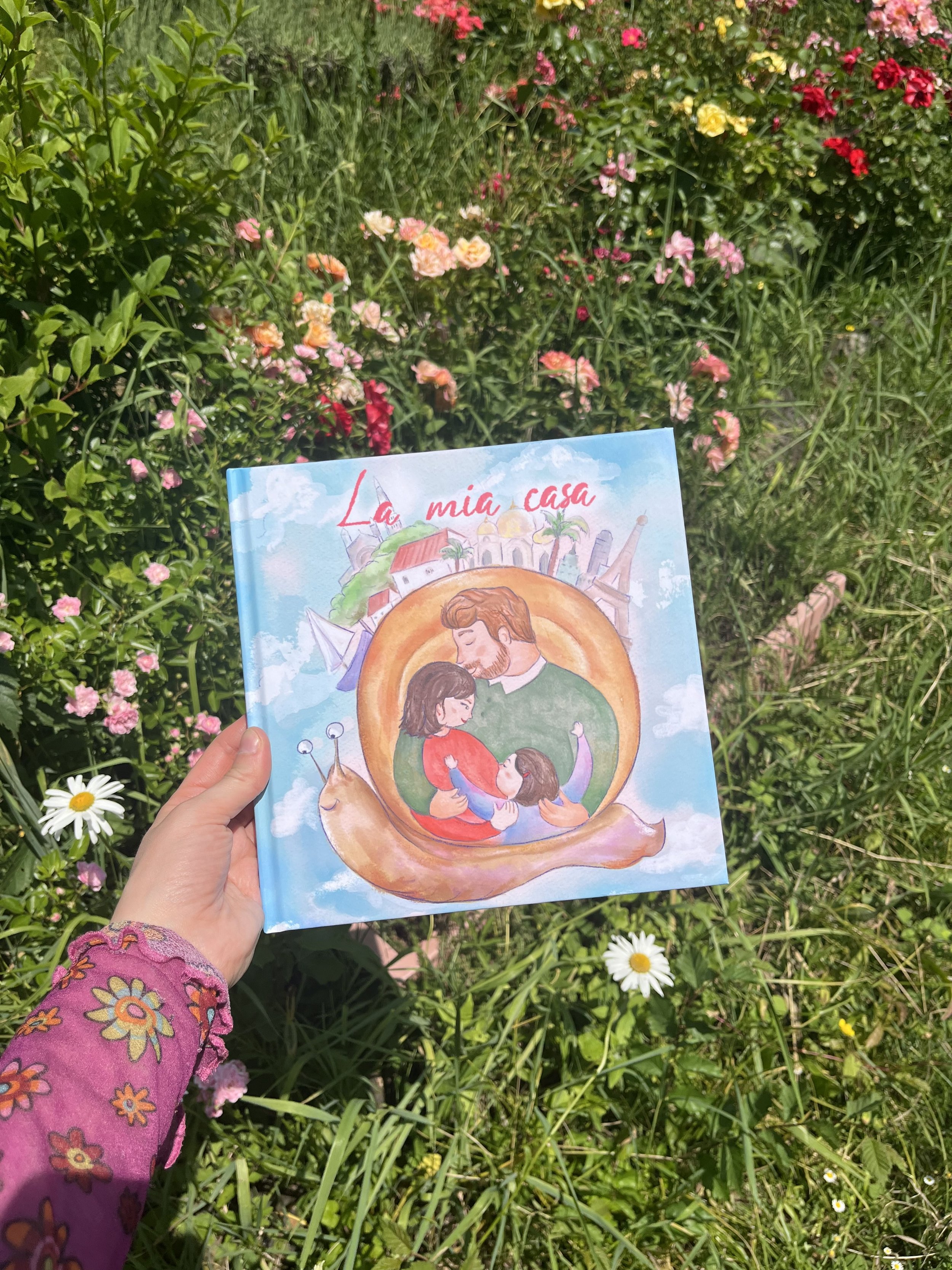 Ragazza che tiene un libro con illustrazione di un abbraccio tra un uomo e una donna in un paesaggio cittadino, con fiori colorati di vario tipo sullo sfondo.