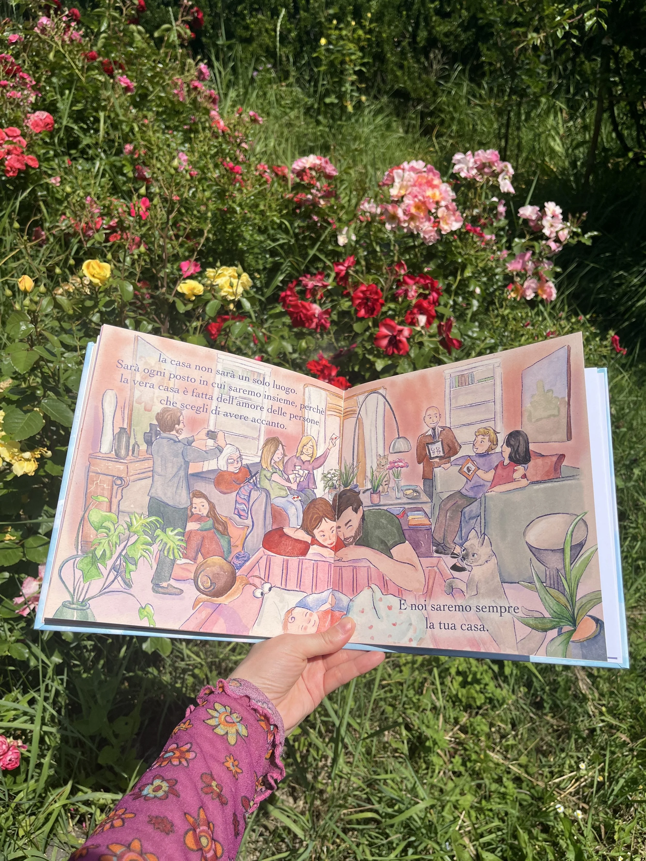 Una persona tiene un libro aperto con illustrazioni di persone che si riuniscono in casa, mentre si trova in un giardino con fiori rosa, rossi e gialli verdi e piante.