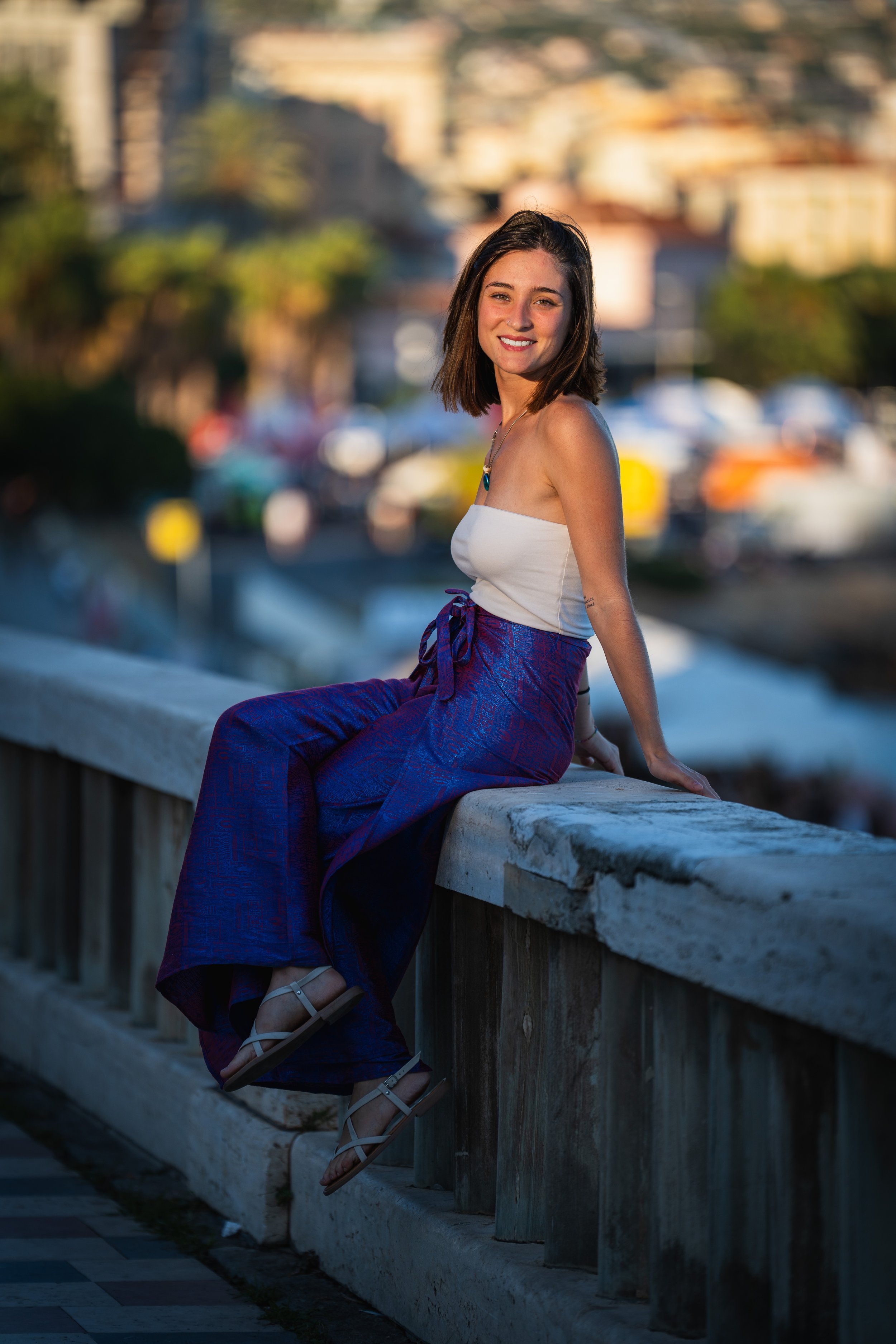 Una giovane donna sorridente seduta sul parapetto di un ponte durante il tramonto, con città e alberi sullo sfondo.