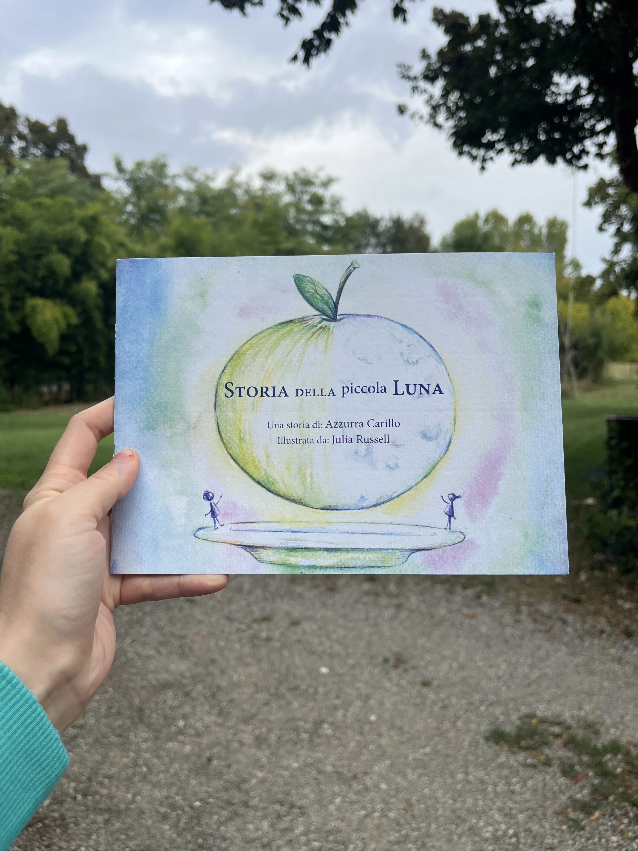 The cover of a book titled “La storia della piccola luna,” featuring a drawing of a stylized apple. The cover is painted in watercolors.