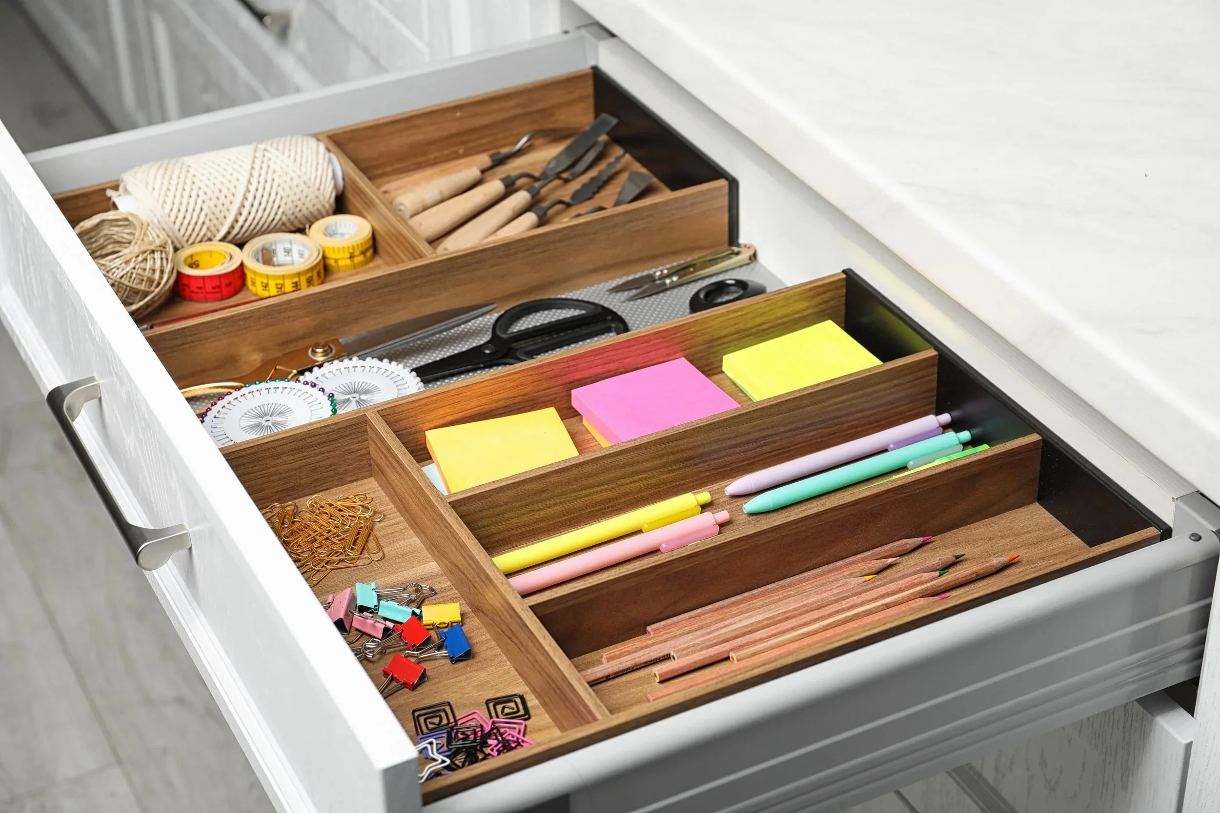 Open drawer with organized compartments containing various stationery items including sticky notes, pens, scissors, tape, paper clips, push pins, and crafting tools.