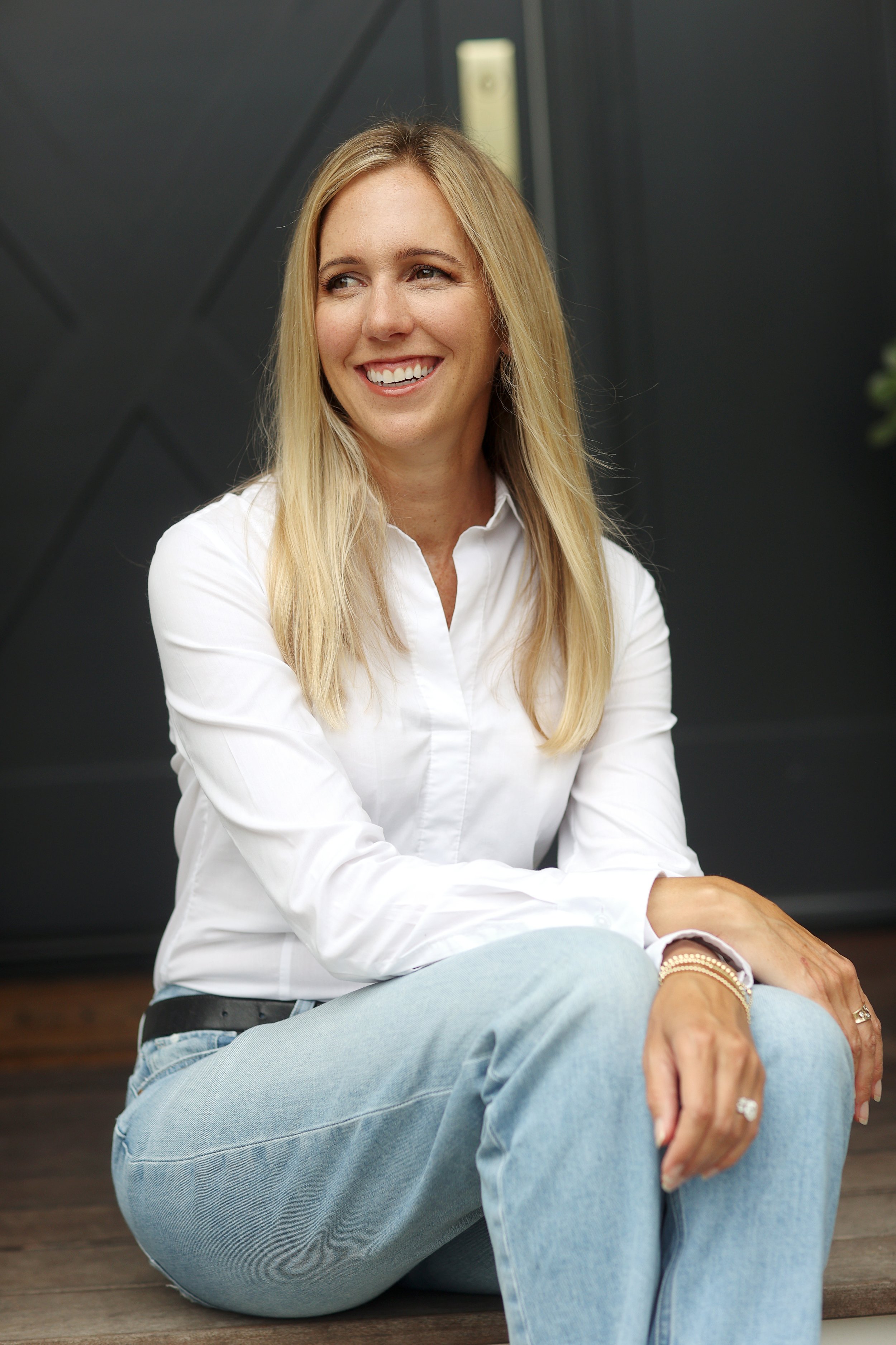 A woman with long blonde hair, wearing a white shirt and light blue jeans, sitting on wooden steps, smiling and looking to the side.