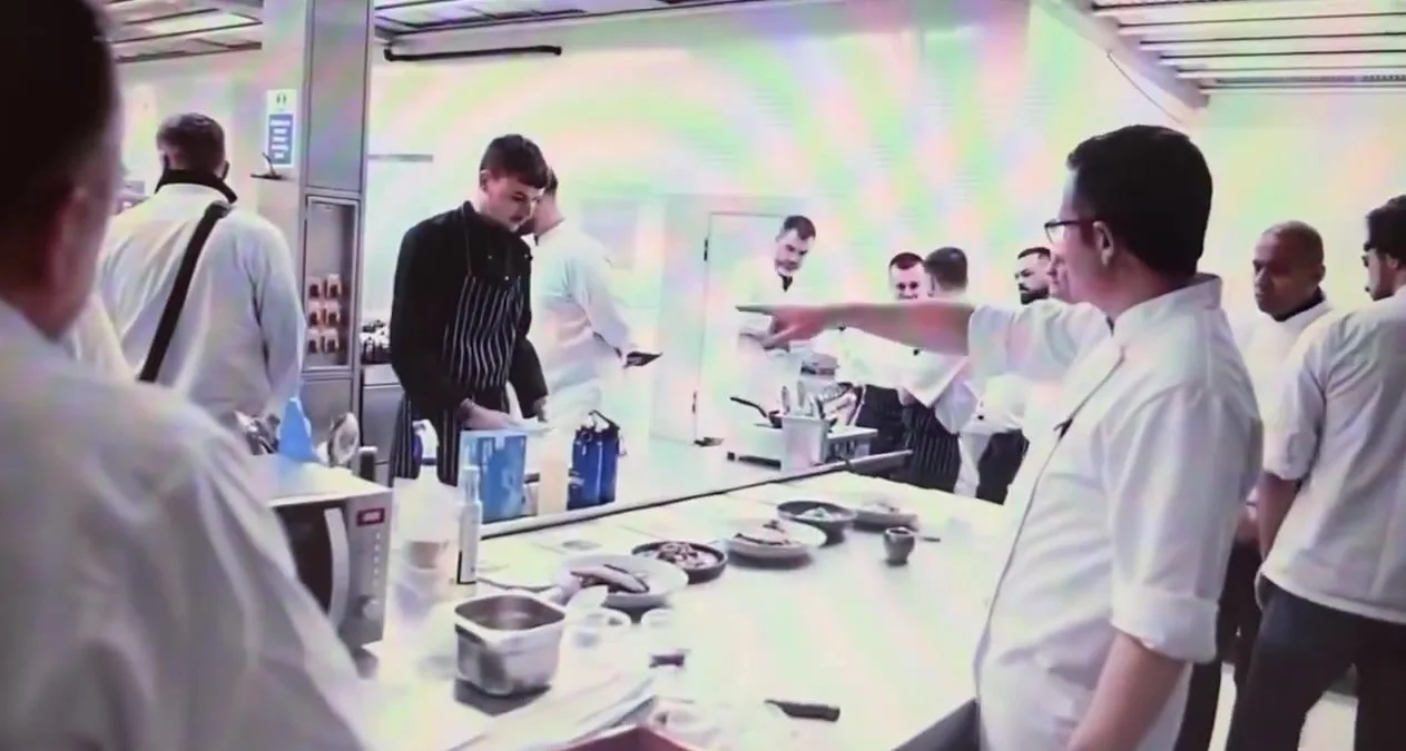 Chefs in whites gathered around a kitchen counter during training