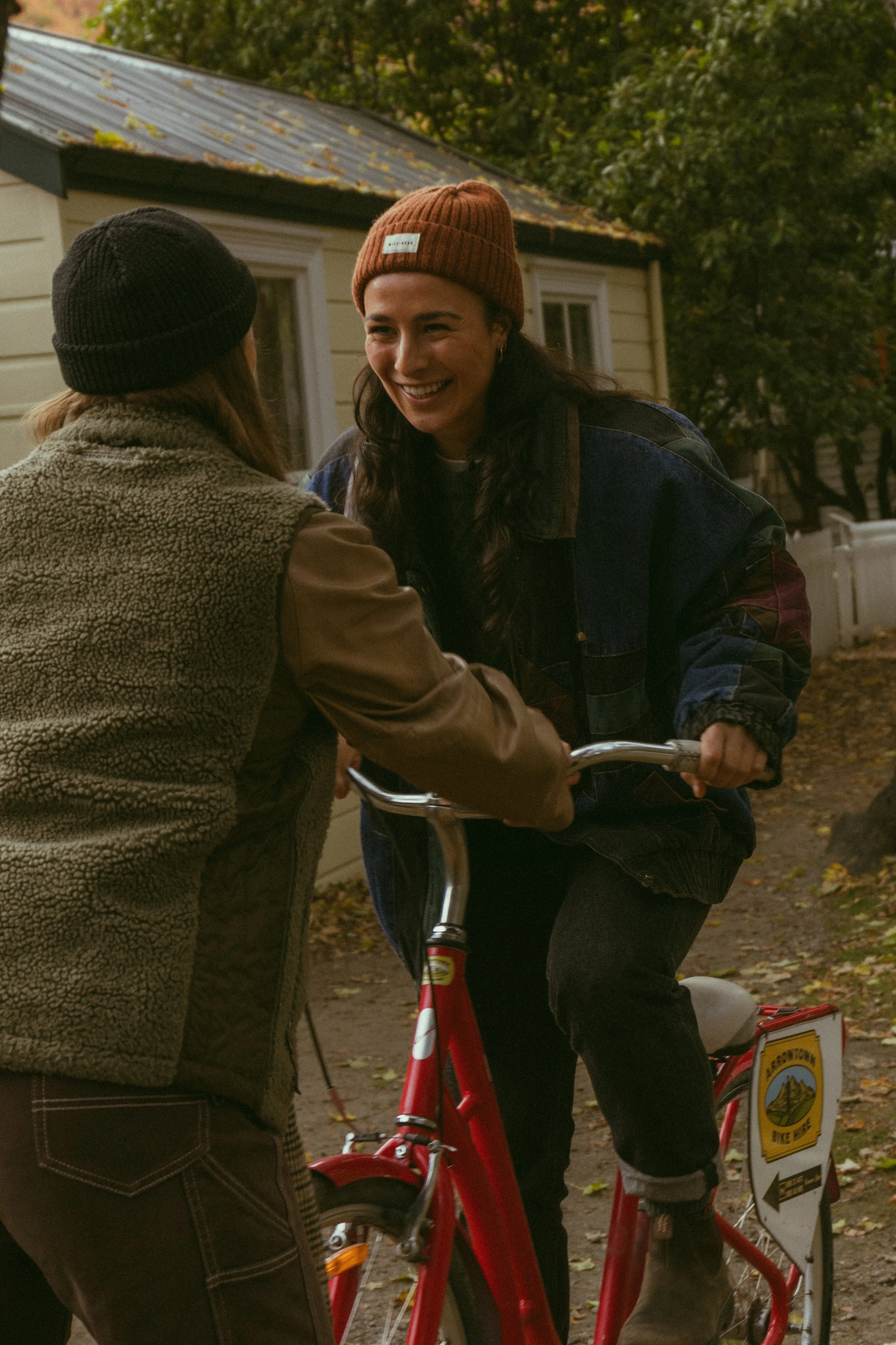 Two women are conversing outdoors on a sidewalk with a yellow house in the background, and one woman is riding a red bicycle. One woman wears a black beanie and a tan jacket with a furry collar, and the other woman is smiling, wearing a brown beanie and a denim jacket. The scene is set during fall with six leaves visible on the ground.