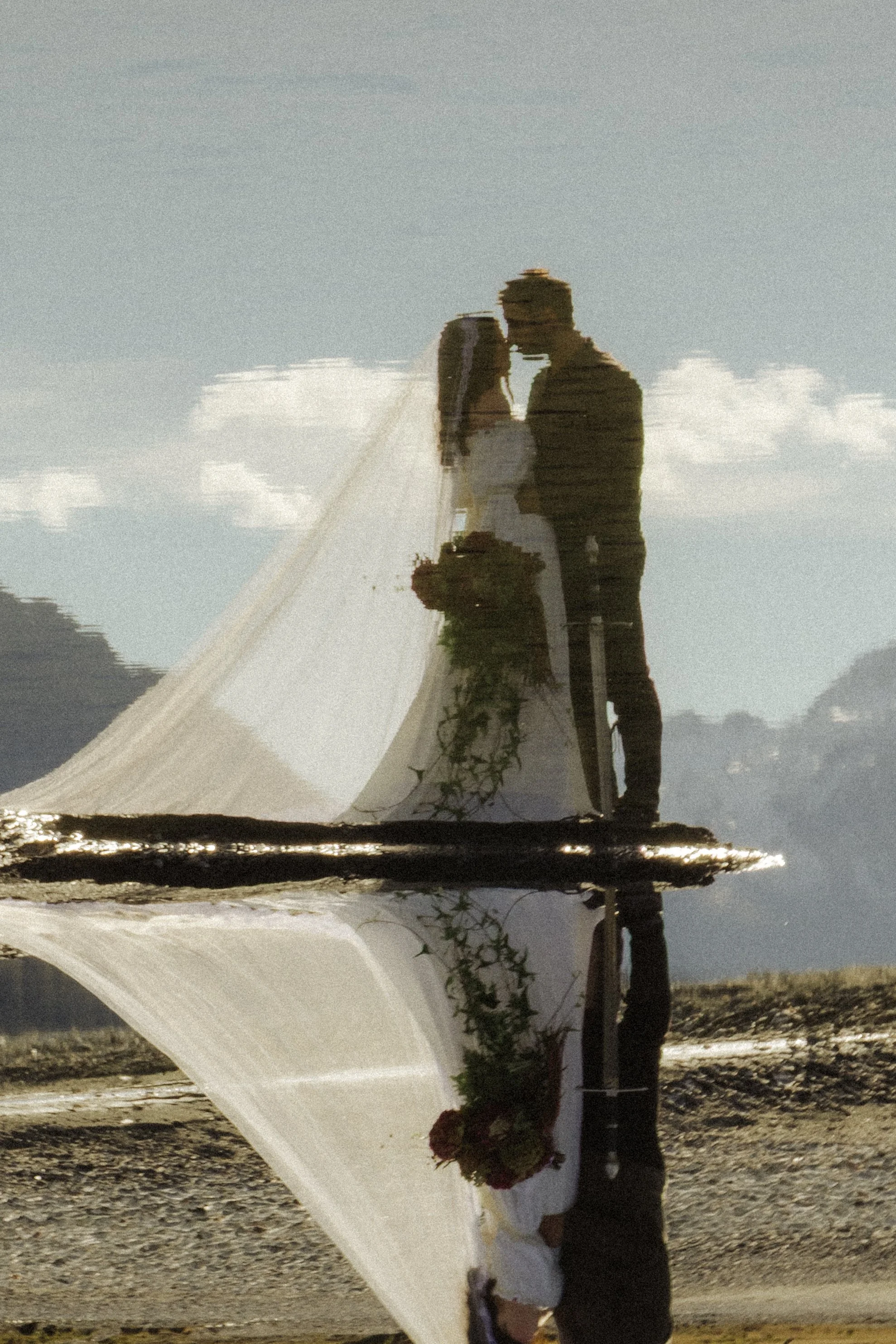 A reflection of a couple holding hands and a bouquet of flowers on a mirror-like surface, with a clear blue sky and some clouds in the background.
