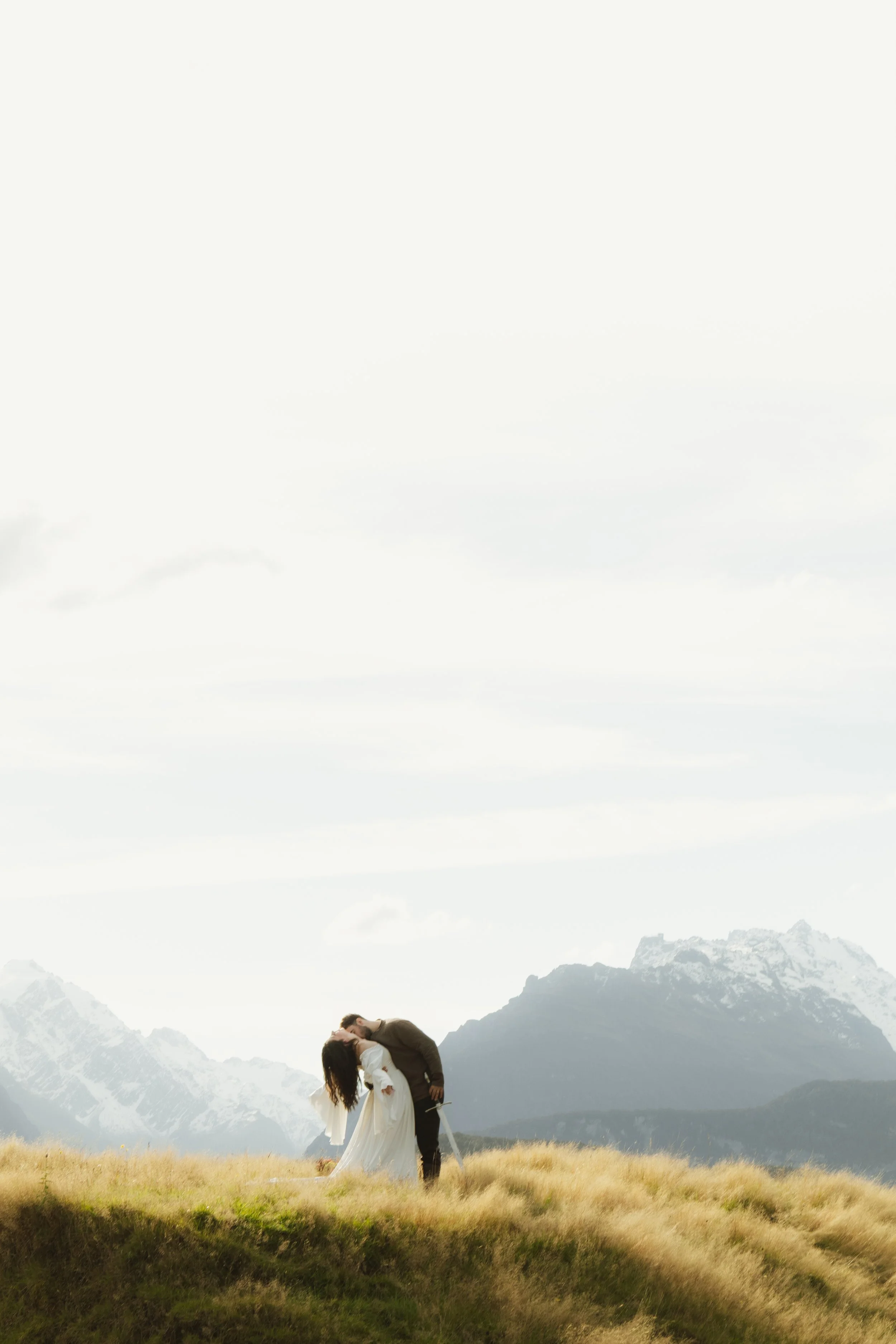 A couple in wedding attire sharing a kiss in a grassy mountain field, with snow-capped peaks and mountains in the background under a cloudy sky.