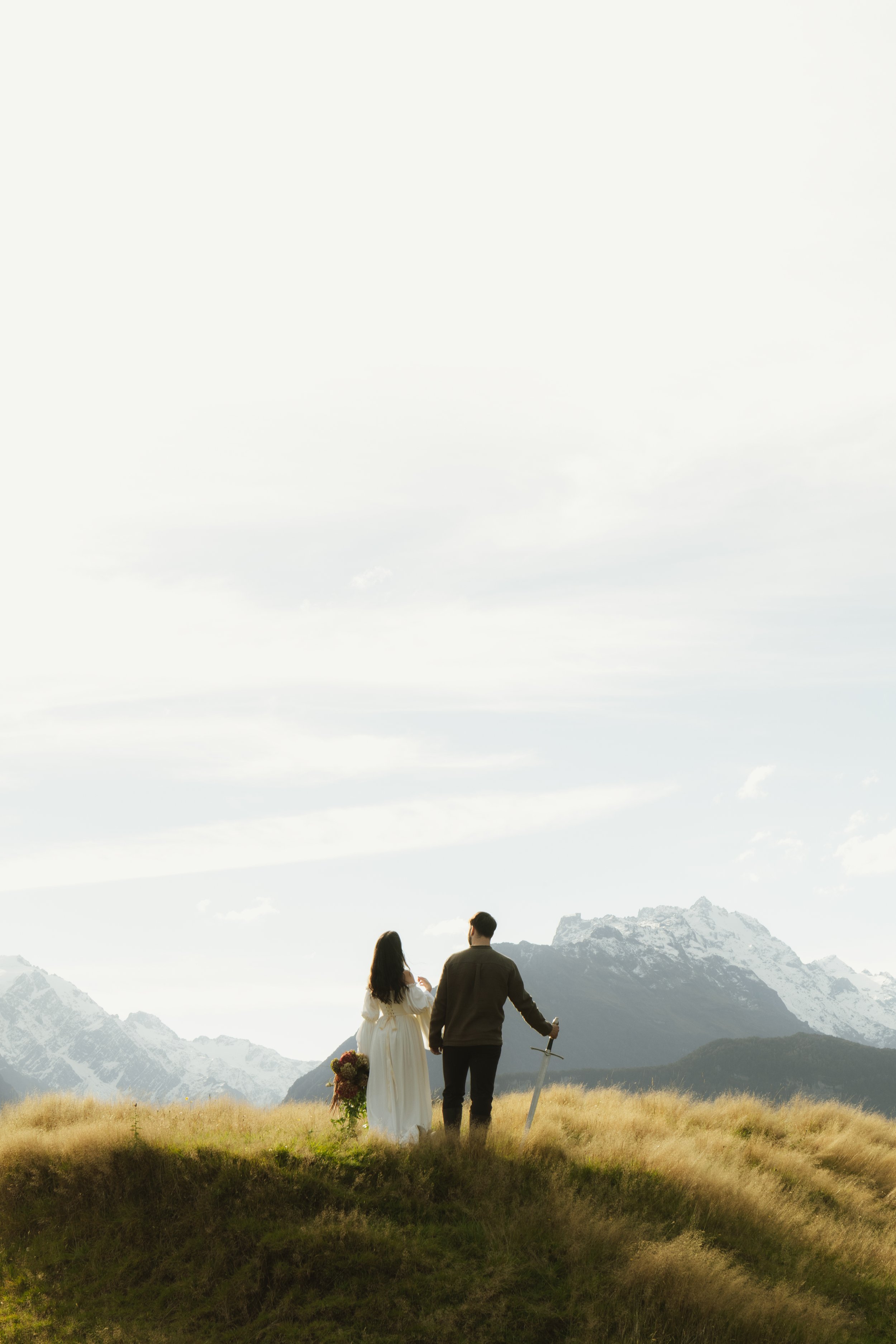 A man and a woman holding hands, walking on a grassy mountainside with snow-capped peaks in the background. The woman wears a white dress and holds a bouquet, while the man is dressed in a dark shirt and pants, holding a sword. The sky is mostly cloudy with patchy sunlight.