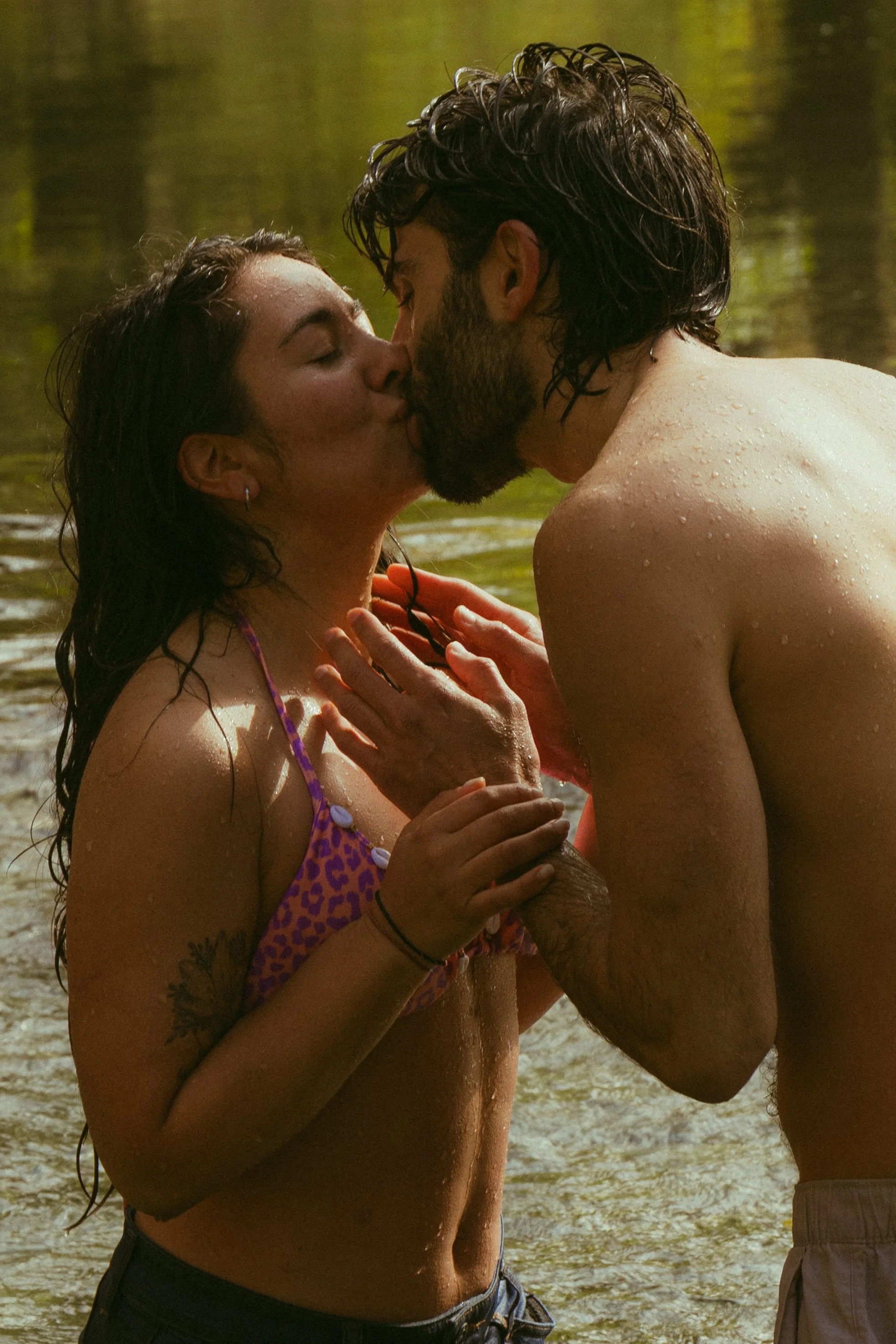 A man and woman kissing in a body of water, with the woman wearing a purple animal print swimsuit and the man shirtless, both wet, with greenery in the background.