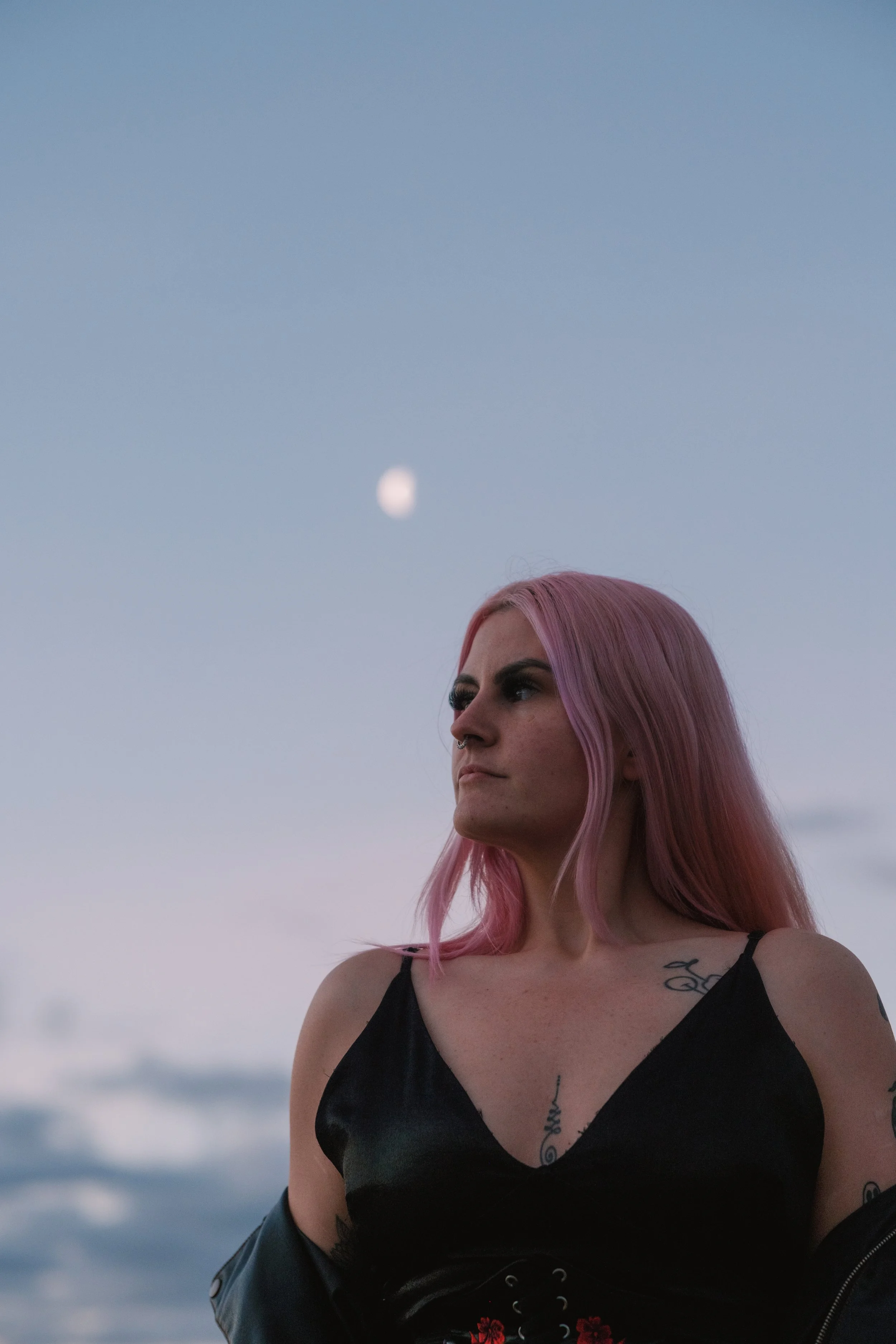 A woman with pink hair wearing a black top and a black jacket, standing outdoors with a cloudy sky and the moon in the background.