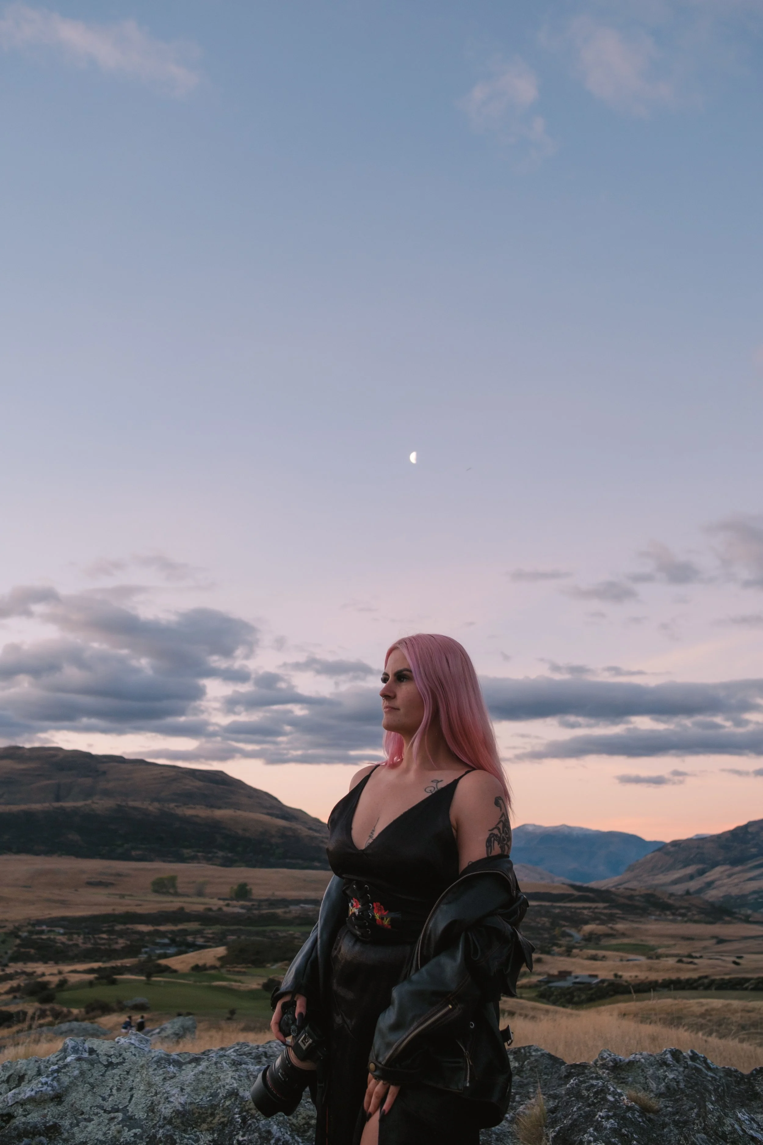 A woman with pink hair wearing a black dress and leather jacket standing outdoors on a rock with a landscape of hills, mountains, and valleys, under a sky with clouds and a visible moon.