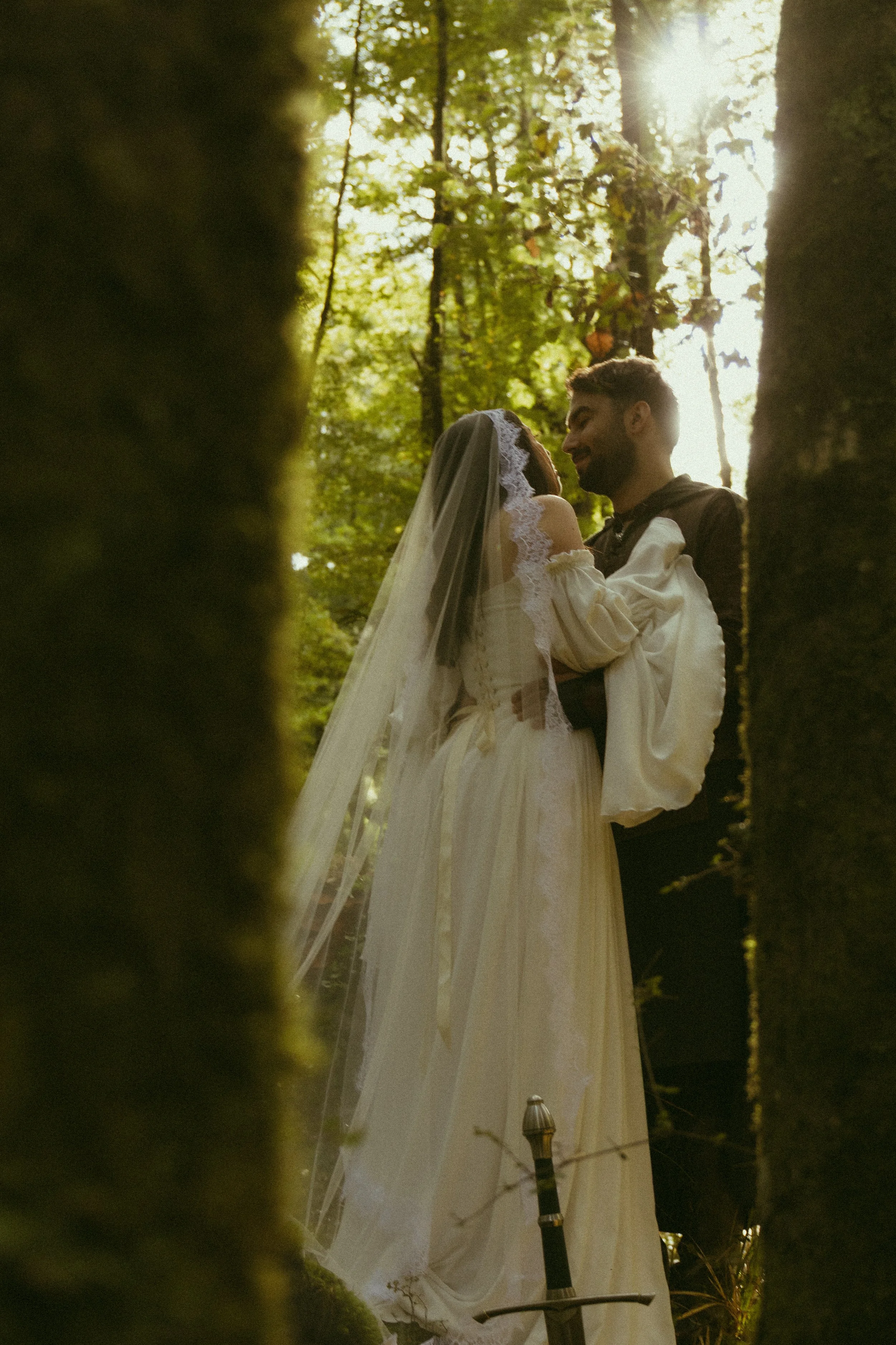 A bride and groom sharing a kiss in a forest, with sunlight filtering through the trees, framed by mossy branches in the foreground.