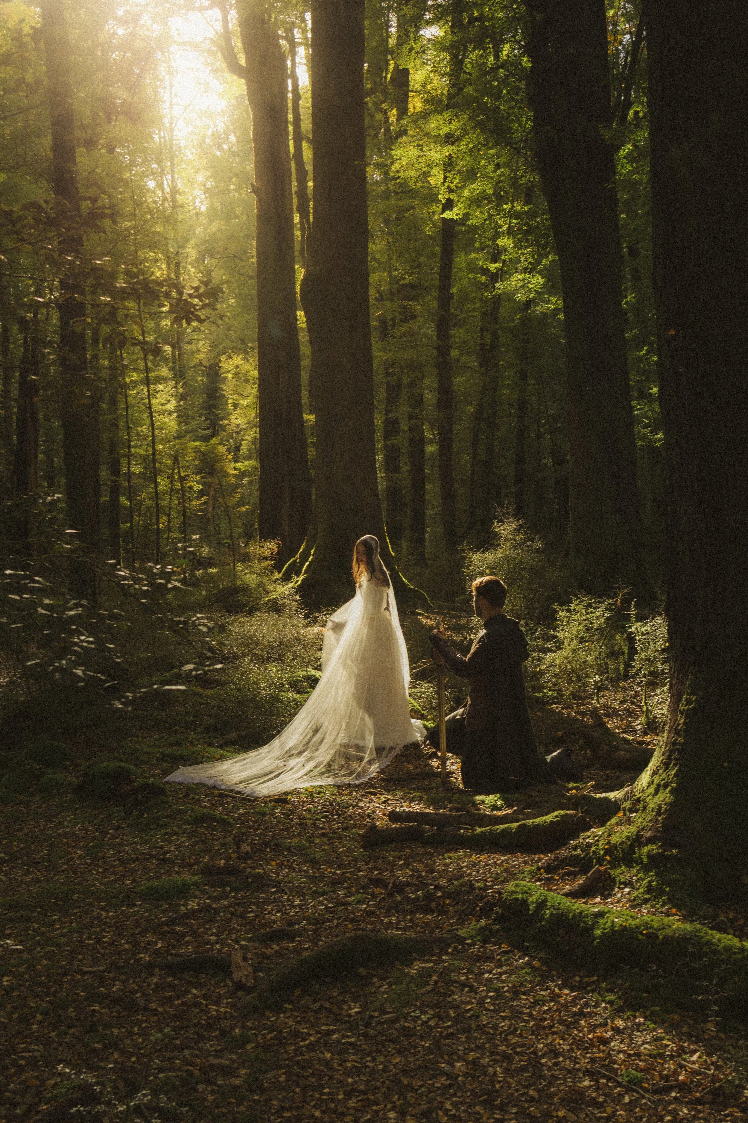 A bride in a white wedding gown and long train standing in a sunlit forest during a wedding ceremony, with an officiant kneeling in front of her, and dressed in dark attire, holding a book or notes. The sunlight filters through the trees, creating a warm, ethereal atmosphere.