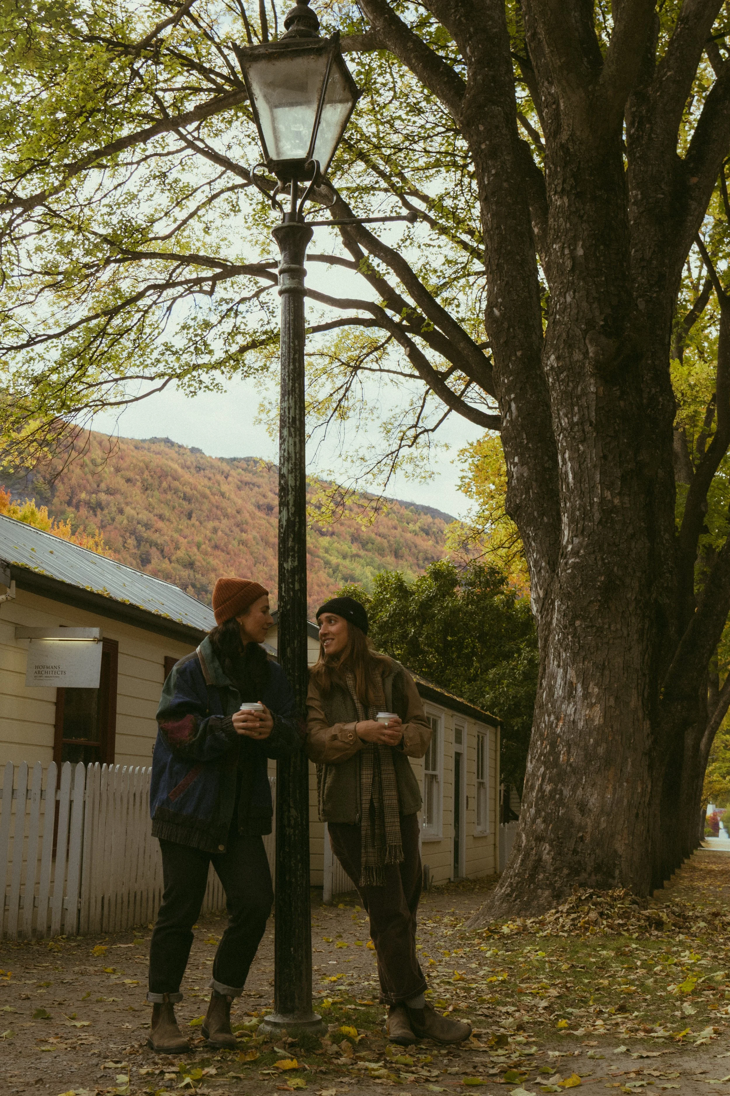 Two women standing outdoors under a large tree, near a vintage street lamp, holding coffee cups, with autumn-colored hills in the background.