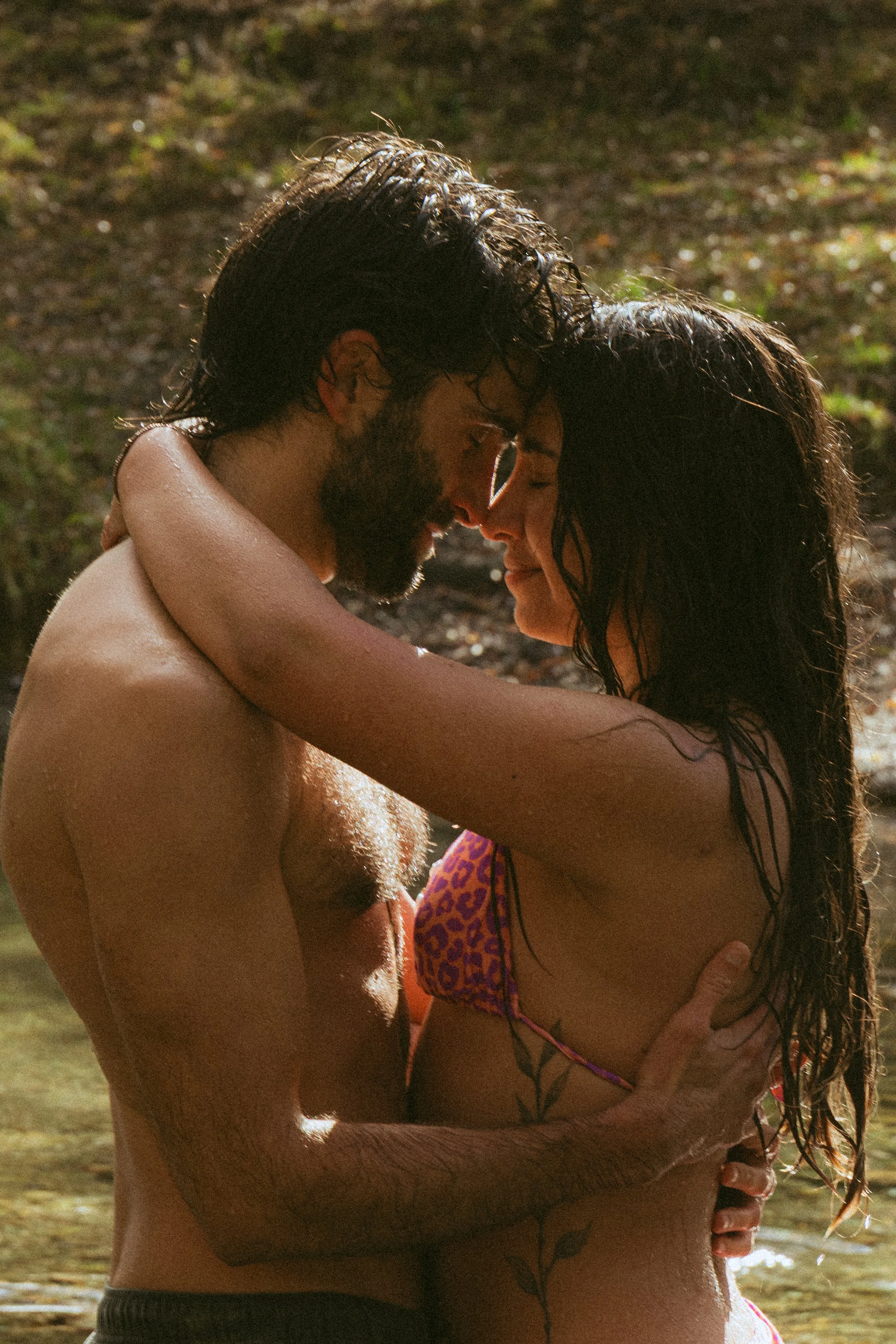 A man and woman embracing each other closely, with their foreheads touching, near a body of water. The man has wet, dark hair, a beard, and is shirtless. The woman has long, wet, dark hair, glasses, and is wearing a leopard print bikini top.