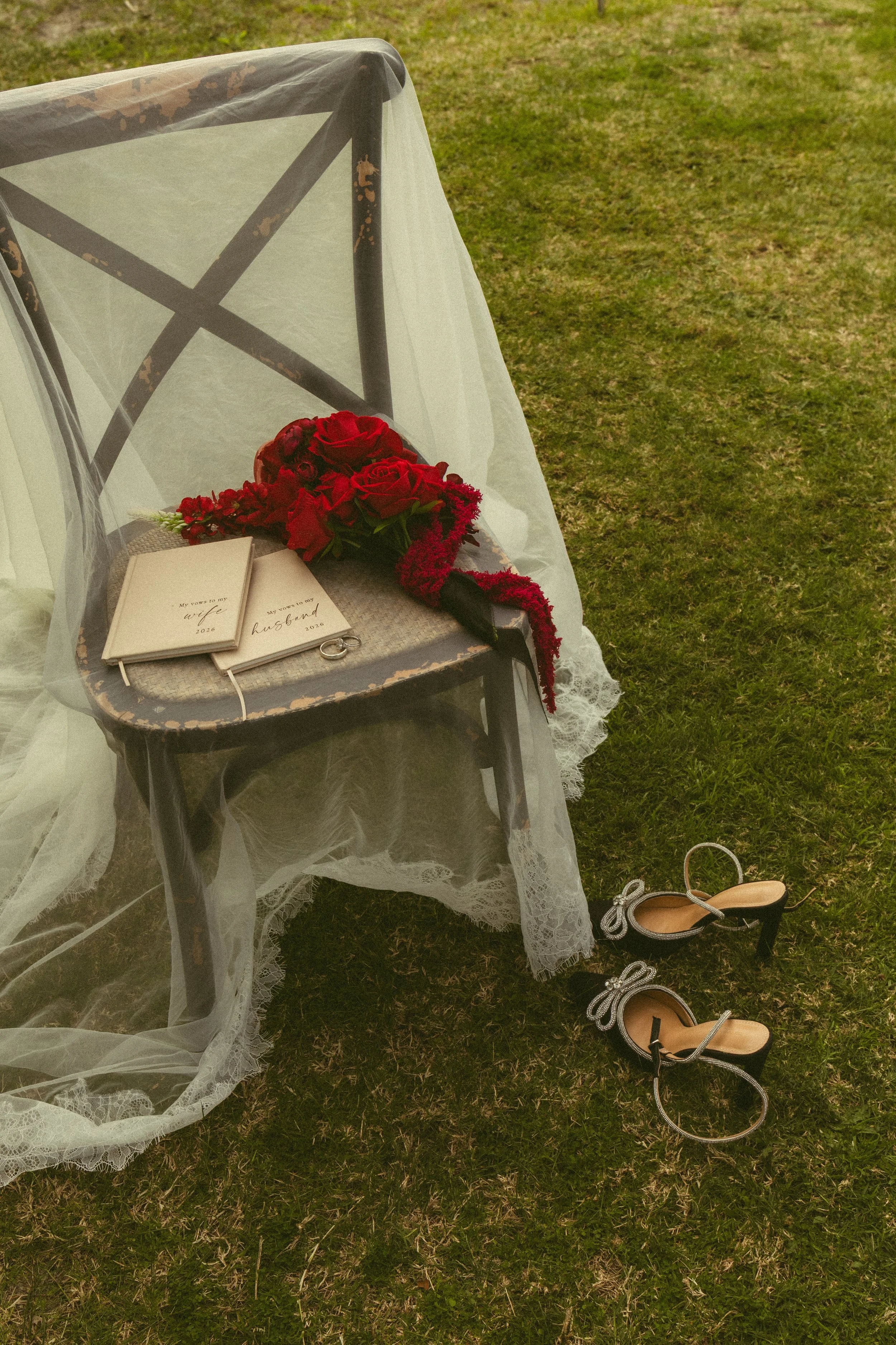 An outdoor scene with a chair draped in white lace fabric, holding a bouquet of red roses and a white book titled "My vow to my wife 2020" and "My vow to my husband 2020." Beside the chair, there are a pair of elegant black and silver high-heeled shoes on the grass.