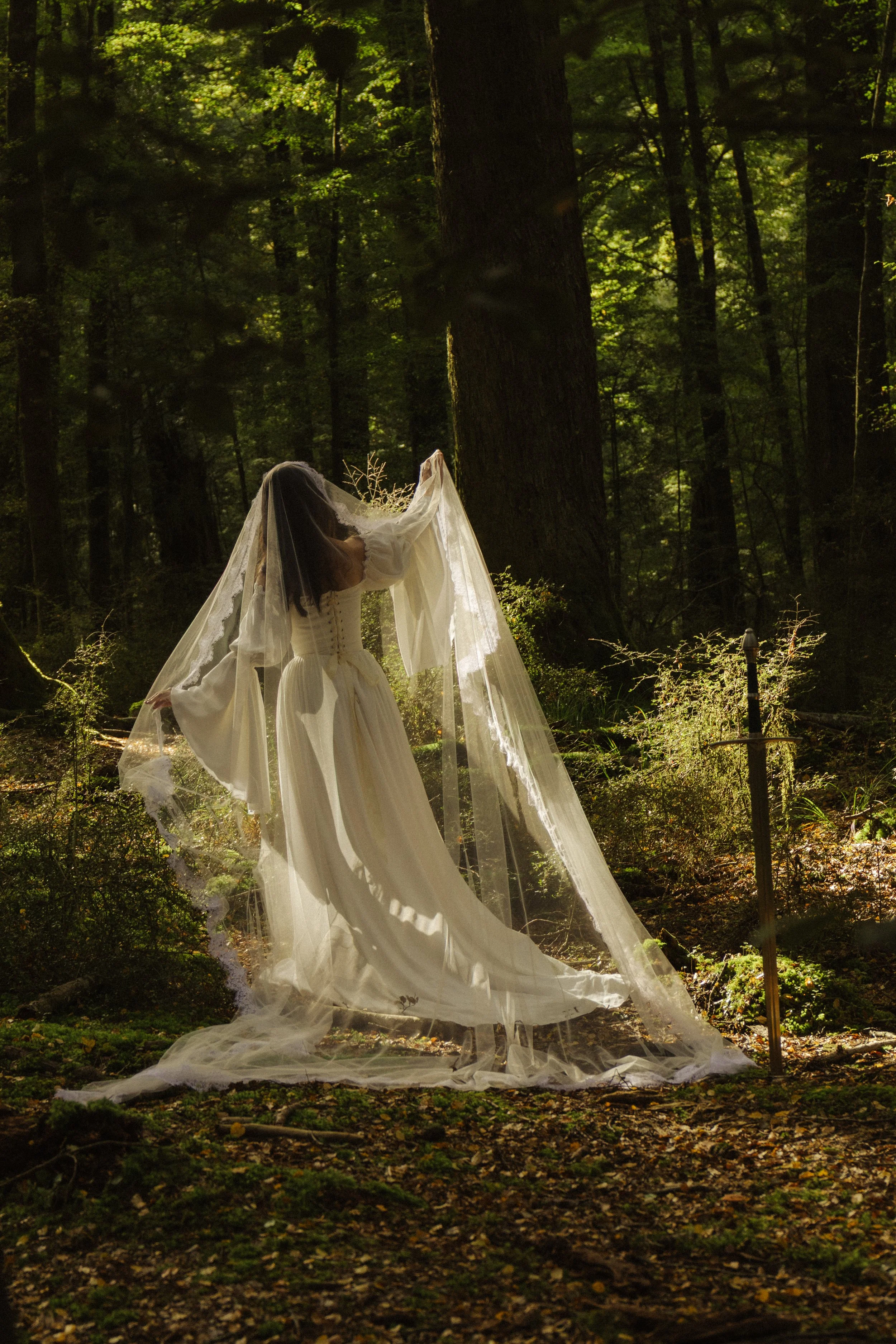 A woman in a vintage white dress with long sleeves and lace details, standing in a forest with tall trees, holding a sheer veil that drapes to the ground, with sunlight filtering through green leaves.