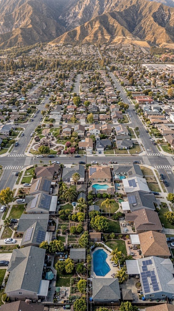Aerial view of a suburban neighborhood with single-family homes, some with swimming pools, surrounded by trees, and mountains in the background.