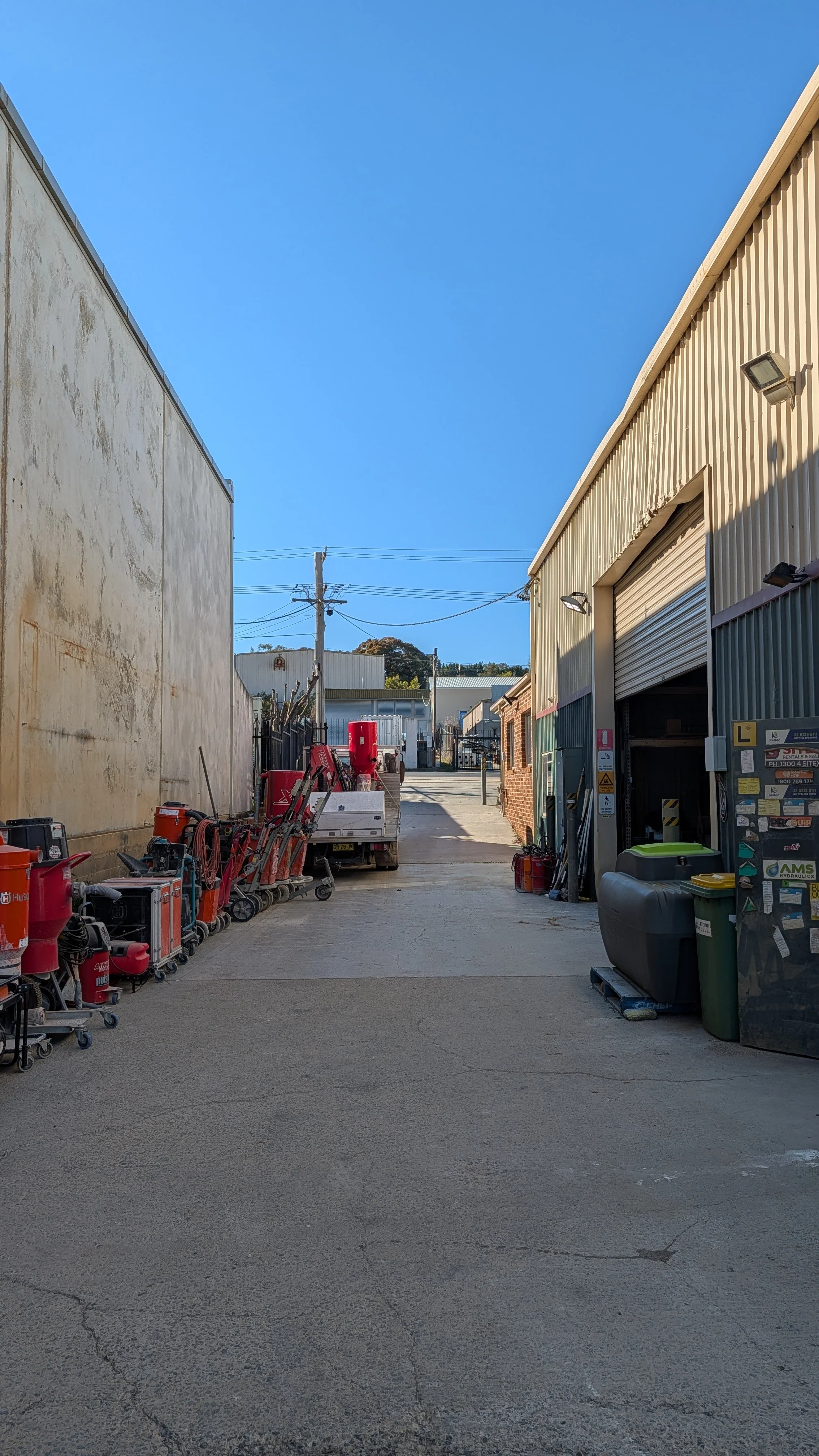 An alleyway between industrial buildings with equipment and tools on either side, utility poles with power lines, and a clear blue sky overhead.