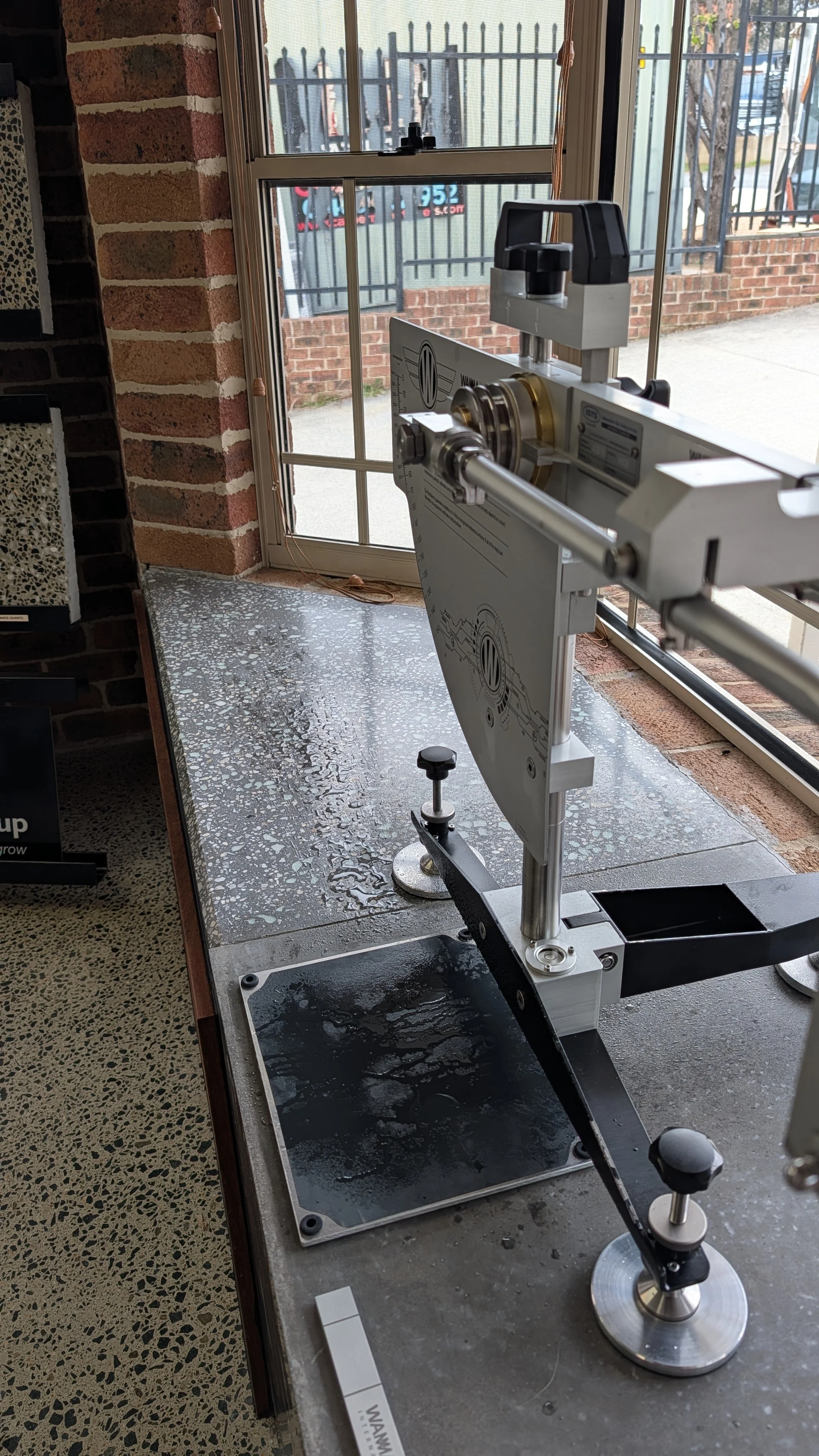 A scientific instrument, likely a spectrometer or precision measurement device, set up near a glass door with a brick wall and fence outside. Water with some condensation is present on the device and table.