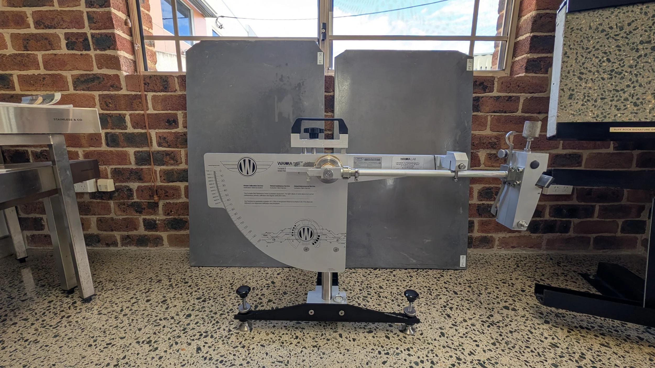 Laboratory equipment with metal surface, measuring and calibration tools, and a brick wall background. The equipment appears to be a testing or calibration device on a speckled floor.