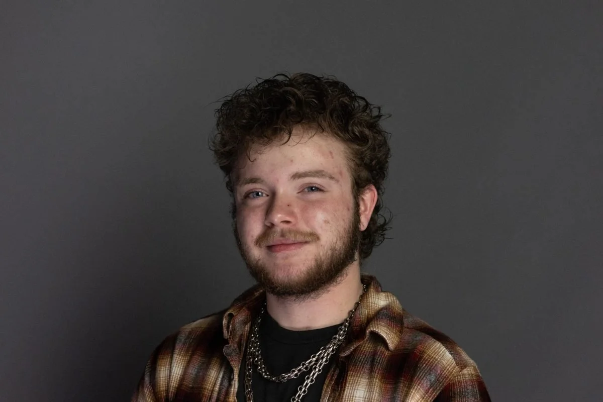 A young man with curly hair, light skin, and a beard, wearing a plaid shirt and layered chains, smiling at the camera against a gray background.