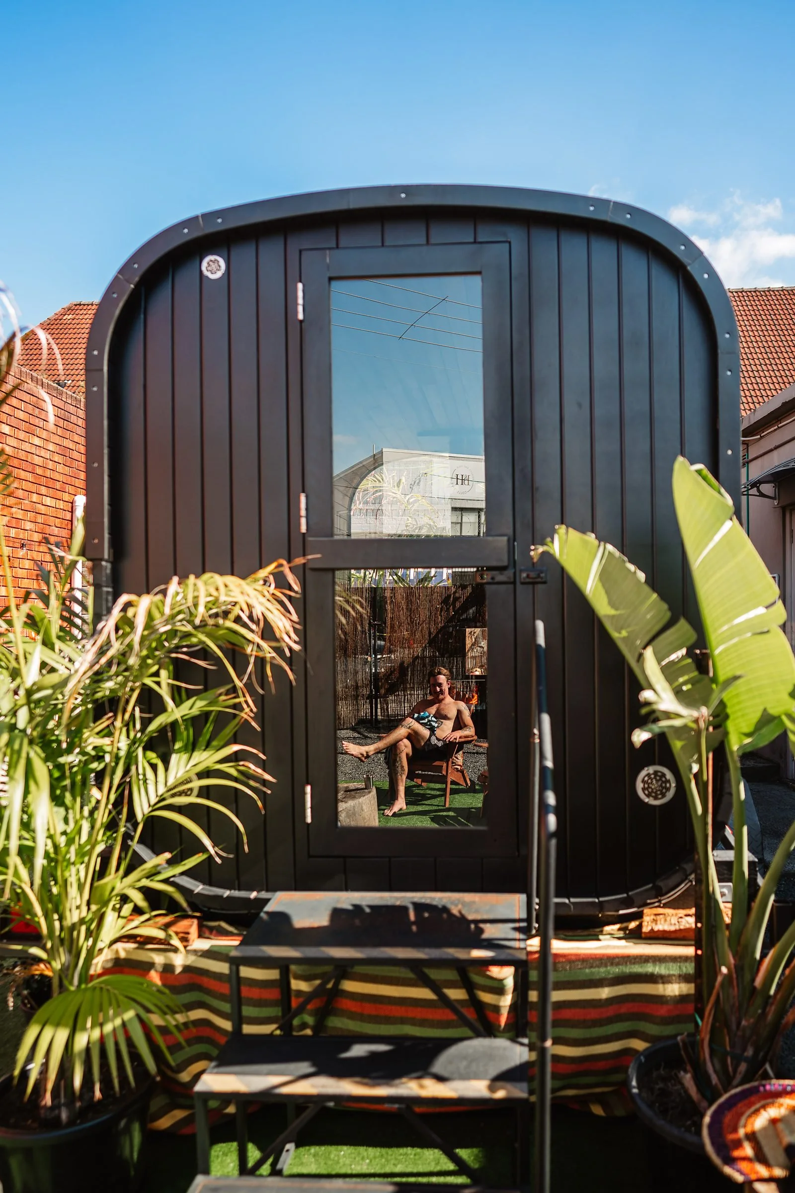 A backyard scene viewed through a black outdoor shower frame, with lounge chairs, plants, and a man relaxing in the background.