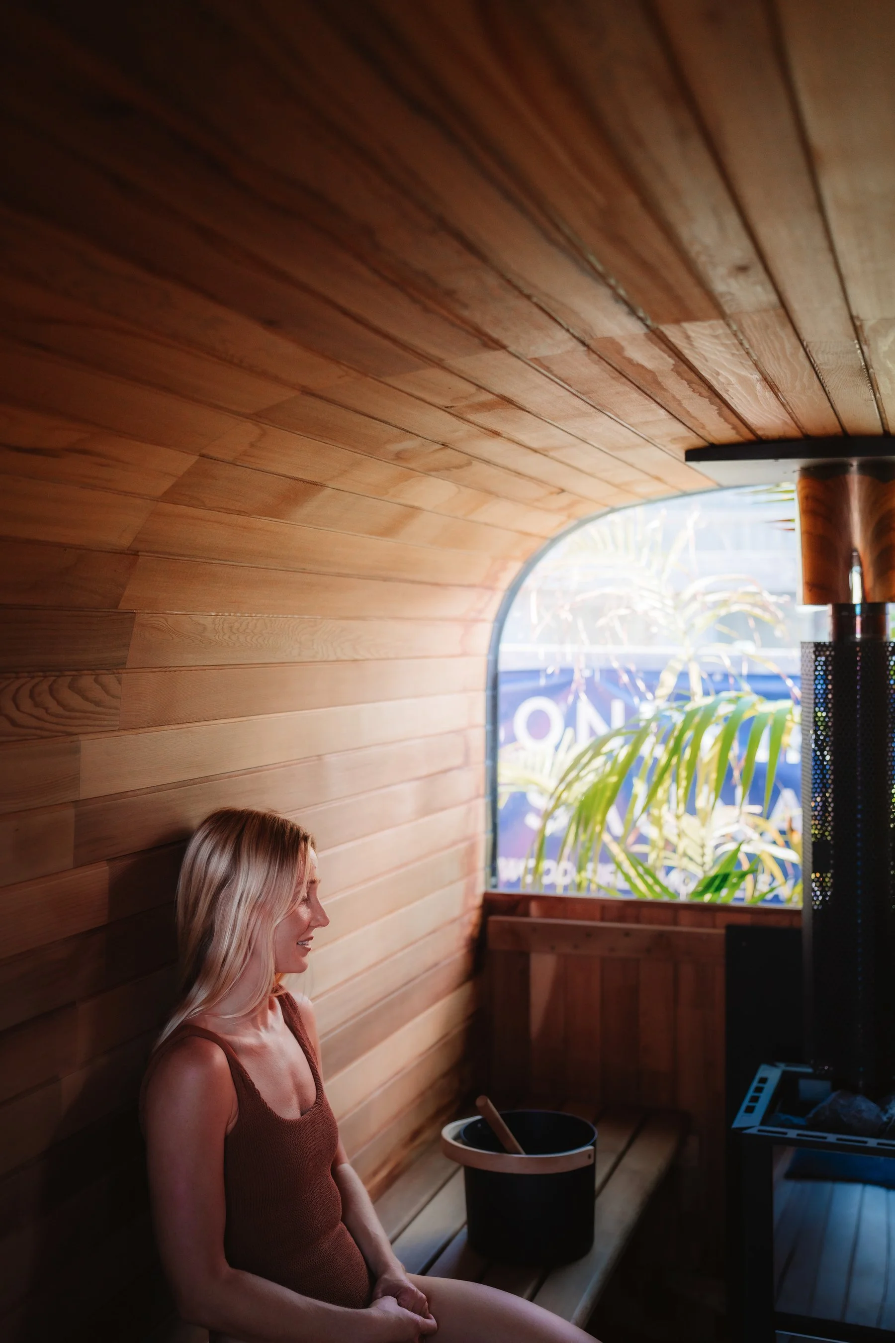 A woman with blonde hair sitting on a bench in a sauna with wooden walls and ceiling, near a window with a view of greenery outside, a bucket with a ladle, and a sauna heater.