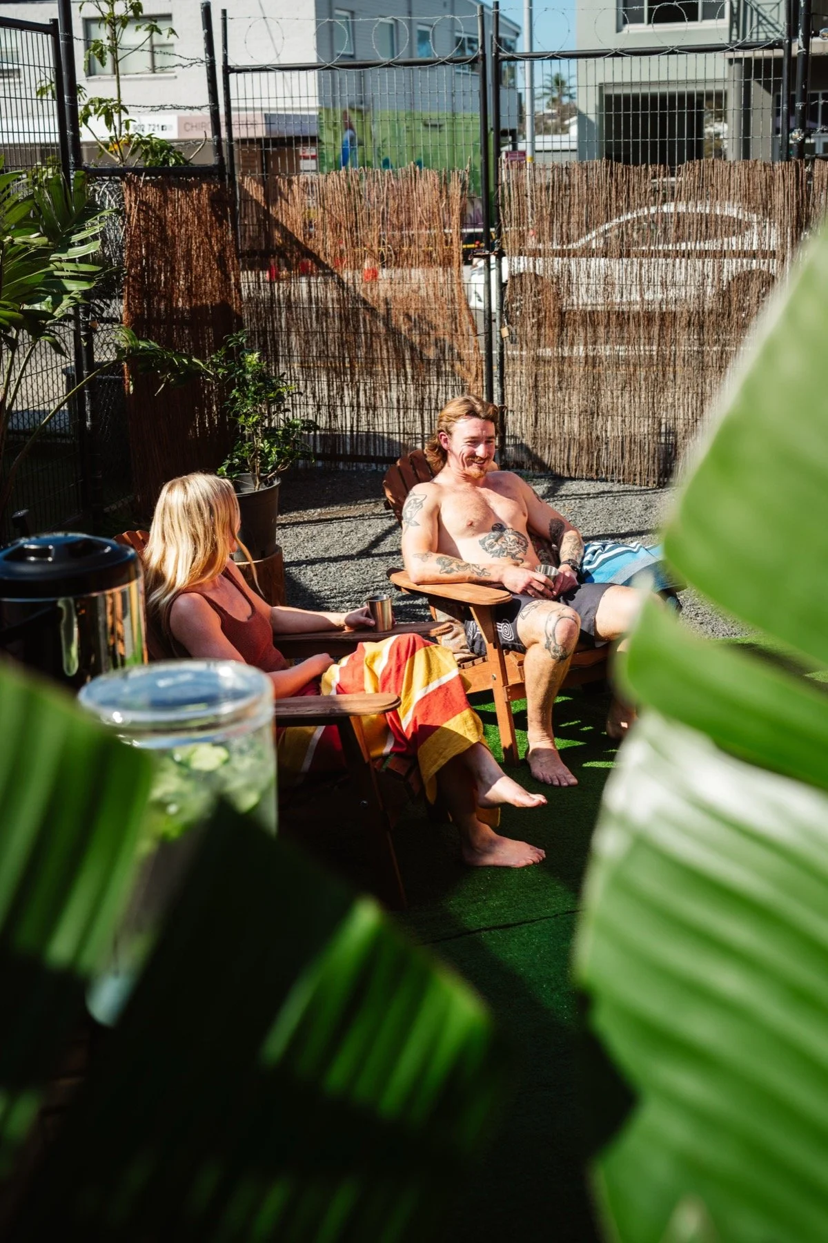 A man with tattoos and long hair relaxing on a wooden chair, smiling, while a woman with blonde hair in a red top and colorful blanket sits next to him on another similar chair. They are outdoors, surrounded by greenery and a fence, with a building and cars in the background.