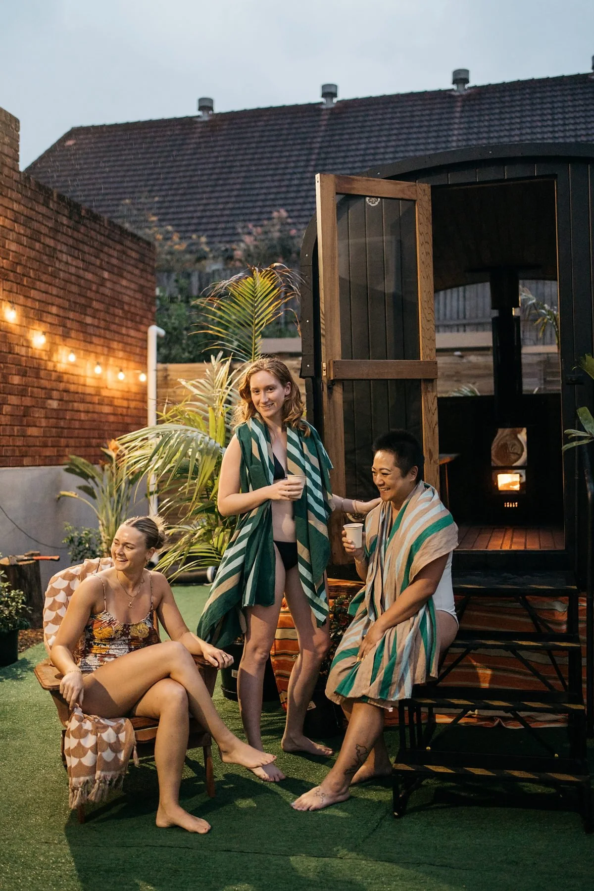 Four women enjoying a backyard gathering in the evening. They are wearing towels or swimsuits, with some holding drinks, sitting and standing on artificial grass near a brick wall decorated with string lights. There are tropical plants, a wooden structure, and a house roof in the background.