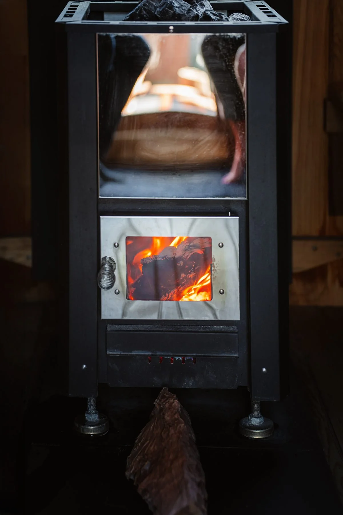 A small black metal wood stove with a glass door burning logs inside, with a piece of wood in front of it.