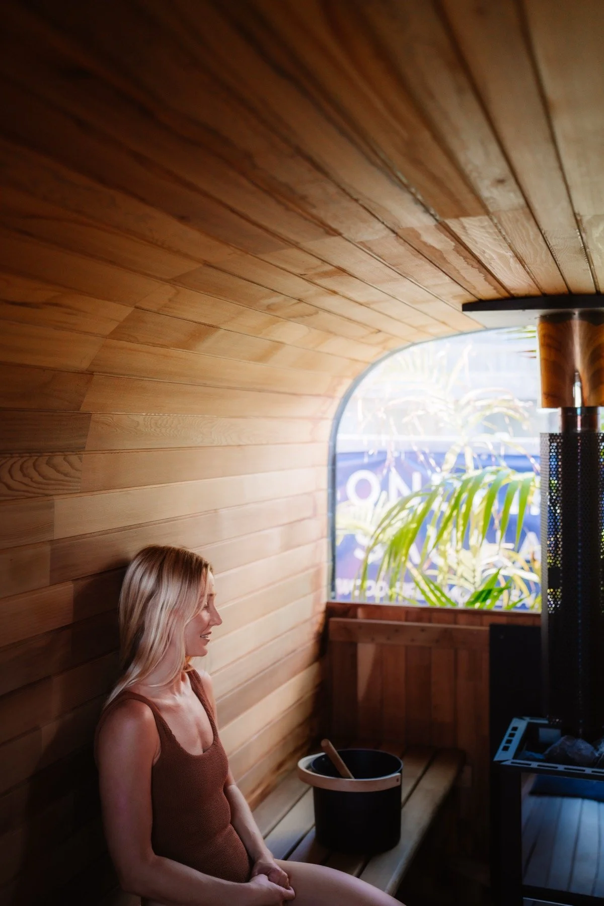 A woman with long blonde hair sitting in a wood-paneled sauna, with a bucket and ladle beside her, near a window showing green plants outside.
