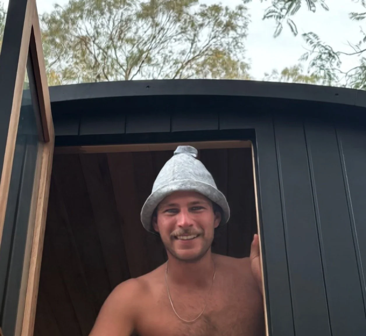 A smiling shirtless man wearing a grey knit hat and a thin gold chain, standing at the open window of a black building with wood interior, outdoors with trees in the background.