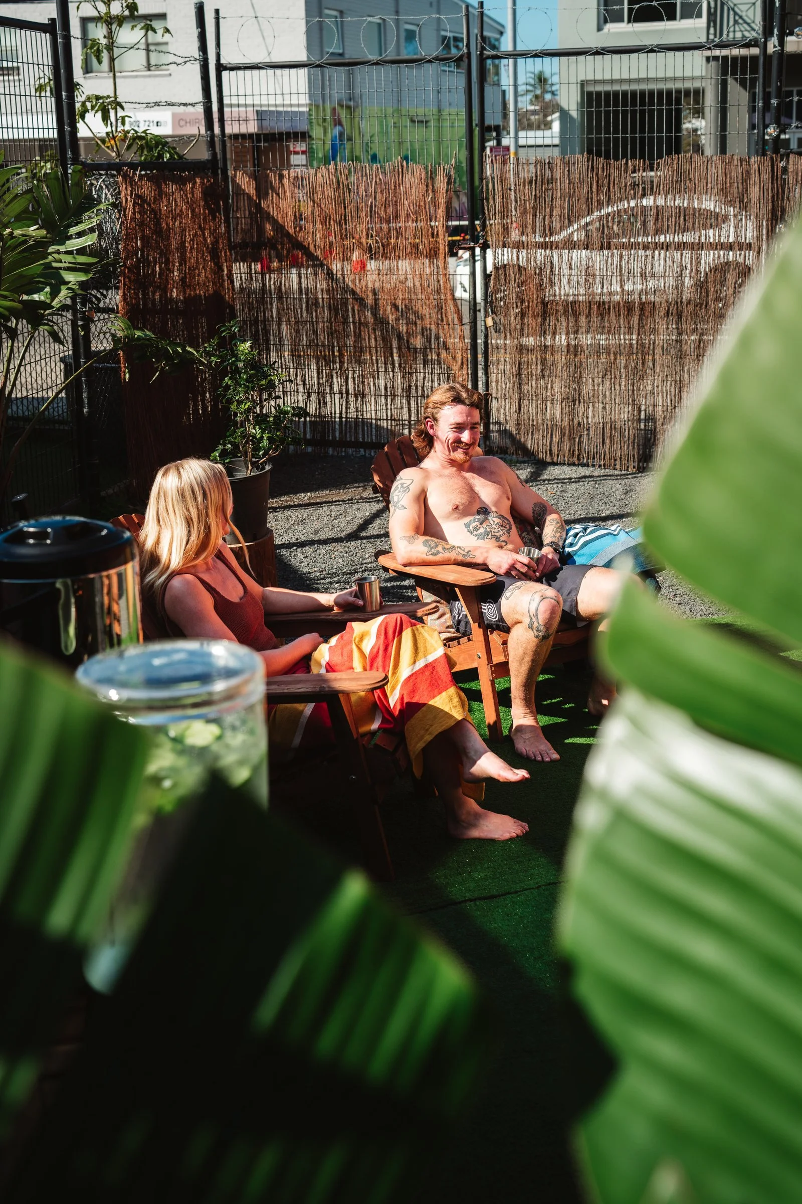 A man with tattoos relaxing outdoors on a lounge chair, with a woman nearby, in a space with green plants, a gravel ground, and a decorative fence with bamboo and wire.
