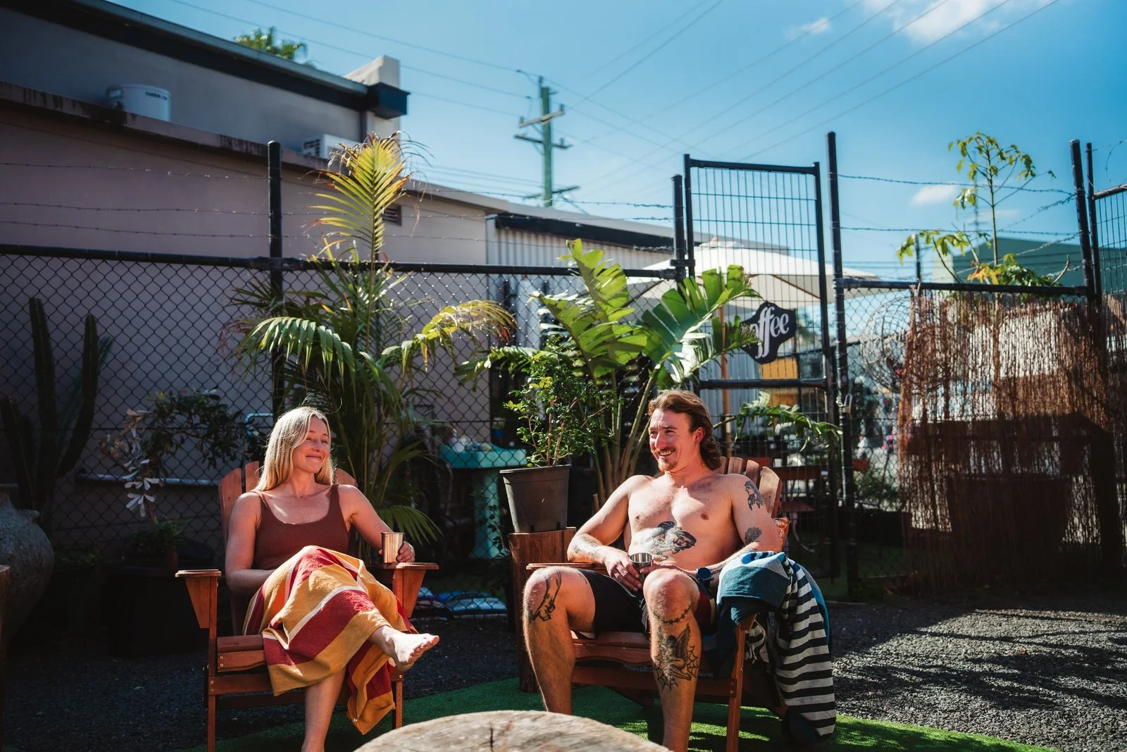 A man and woman sitting outside in Adirondack chairs, smiling and relaxing on a sunny day. The woman has blonde hair, is wearing a brown tank top, and has a colorful towel on her legs. The man has tattoos, dark hair, and is shirtless, with a striped towel on his lap. They are surrounded by plants, with a metal fence and a building in the background.