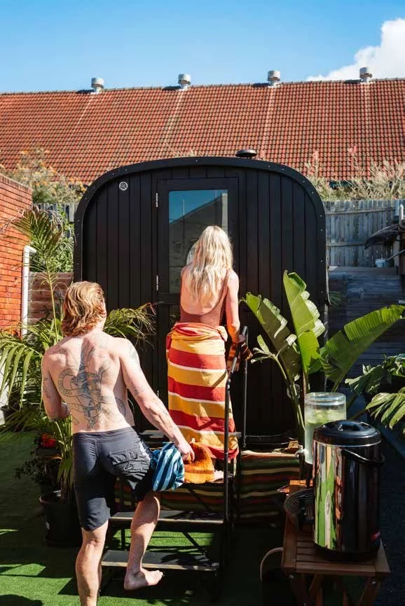 A woman with long blonde hair wrapped in a striped towel is entering a black outdoor shower with a curved roof, in a backyard with tropical plants, while a shirtless man with tattoos and longer hair stands nearby holding a towel, with a bench and a trash can in the foreground.