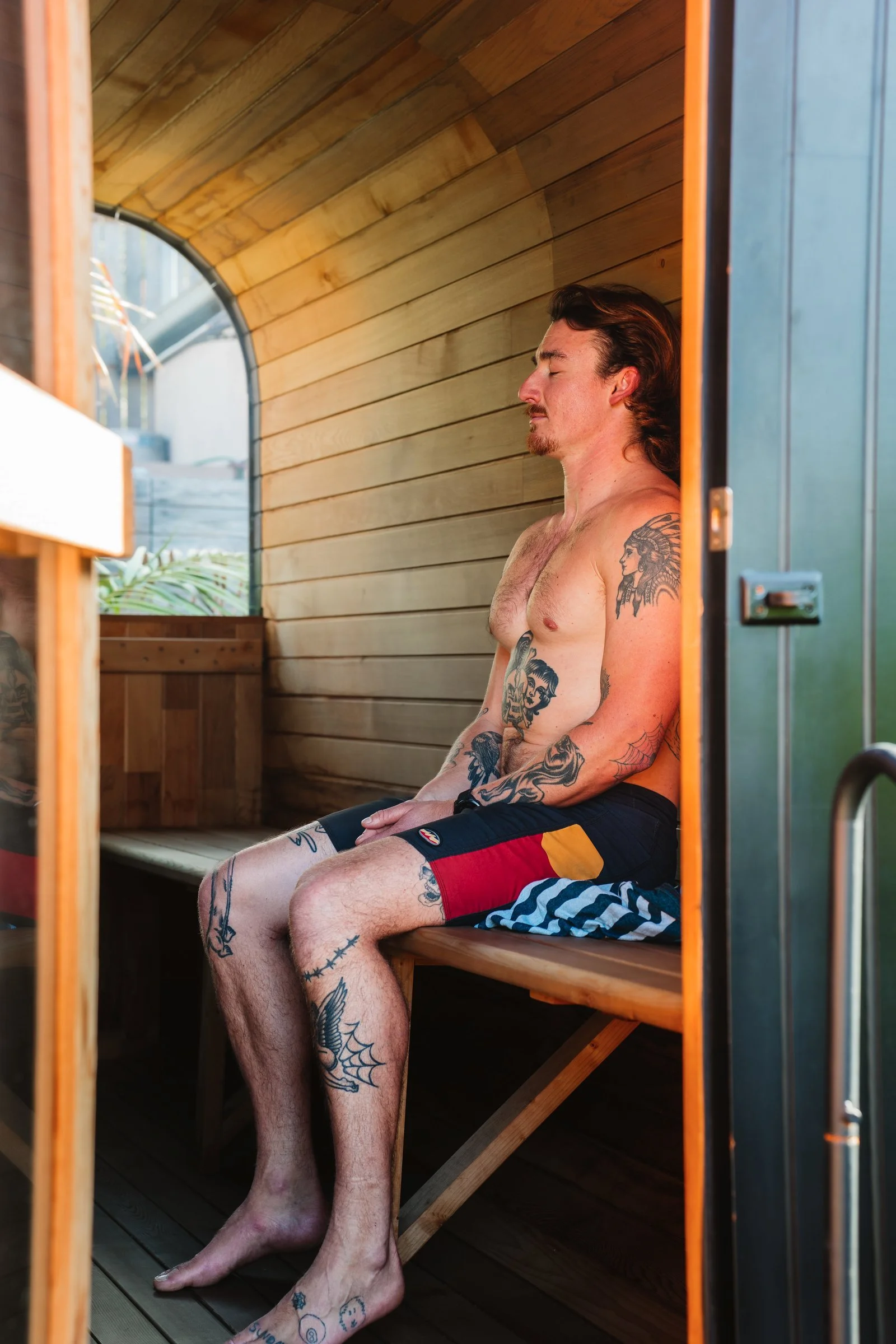 A shirtless man with tattoos sitting inside an outdoor wooden sauna or steam room, with his eyes closed and hands resting on his lap.