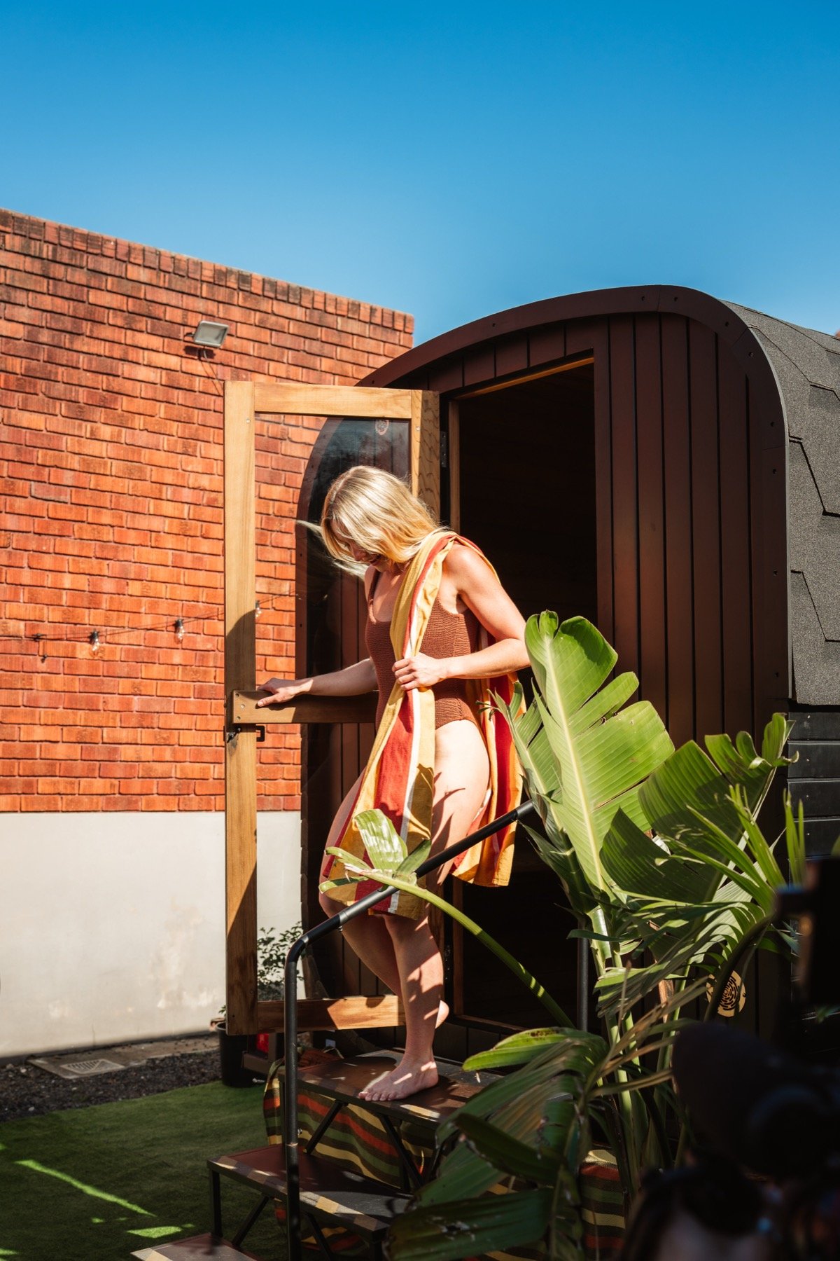 A woman standing on a small wooden platform outside a building with a red brick wall, wearing a brown crop top and a multicolored towel draped over her shoulders, next to lush green plants on a sunny day.