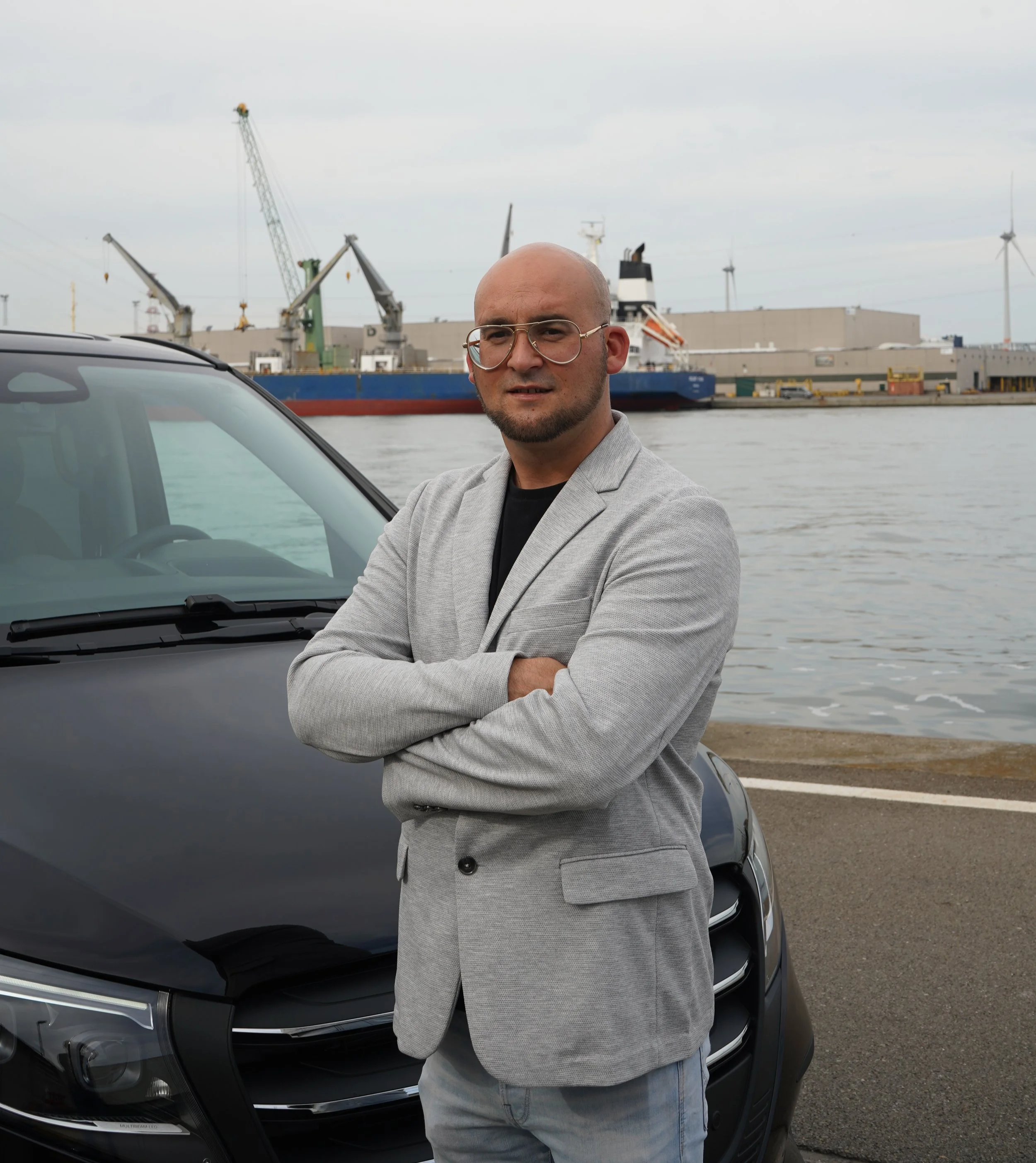Chauffeur van GDS Trans Service poseert bij een zwarte auto aan het water, met een industriële haven, kranen en windturbines op de achtergrond.