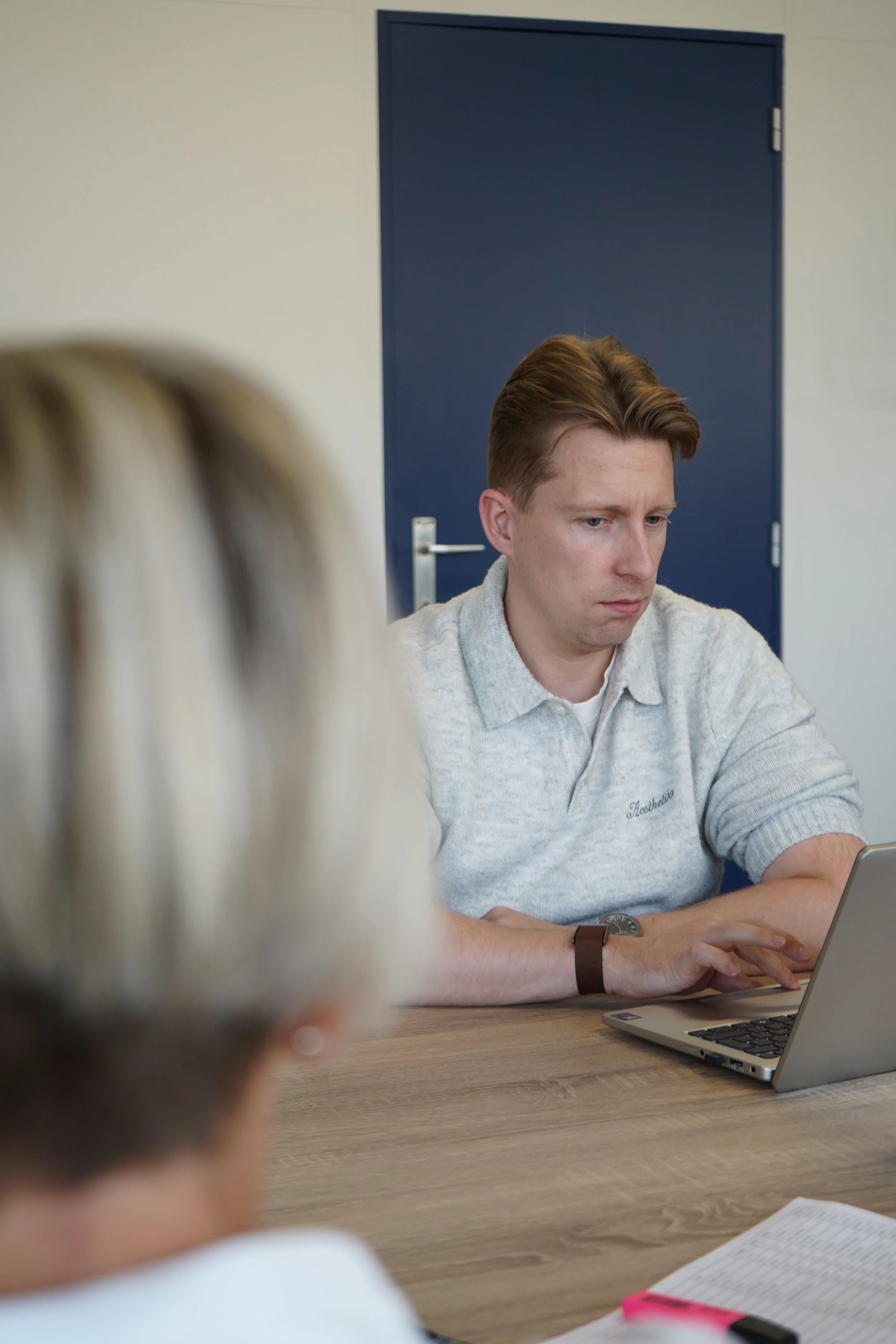 Andy van GDS Trans Service in lichtgrijs shirt werkt op een laptop aan een houten tafel, met een blauwe deur op de achtergrond.